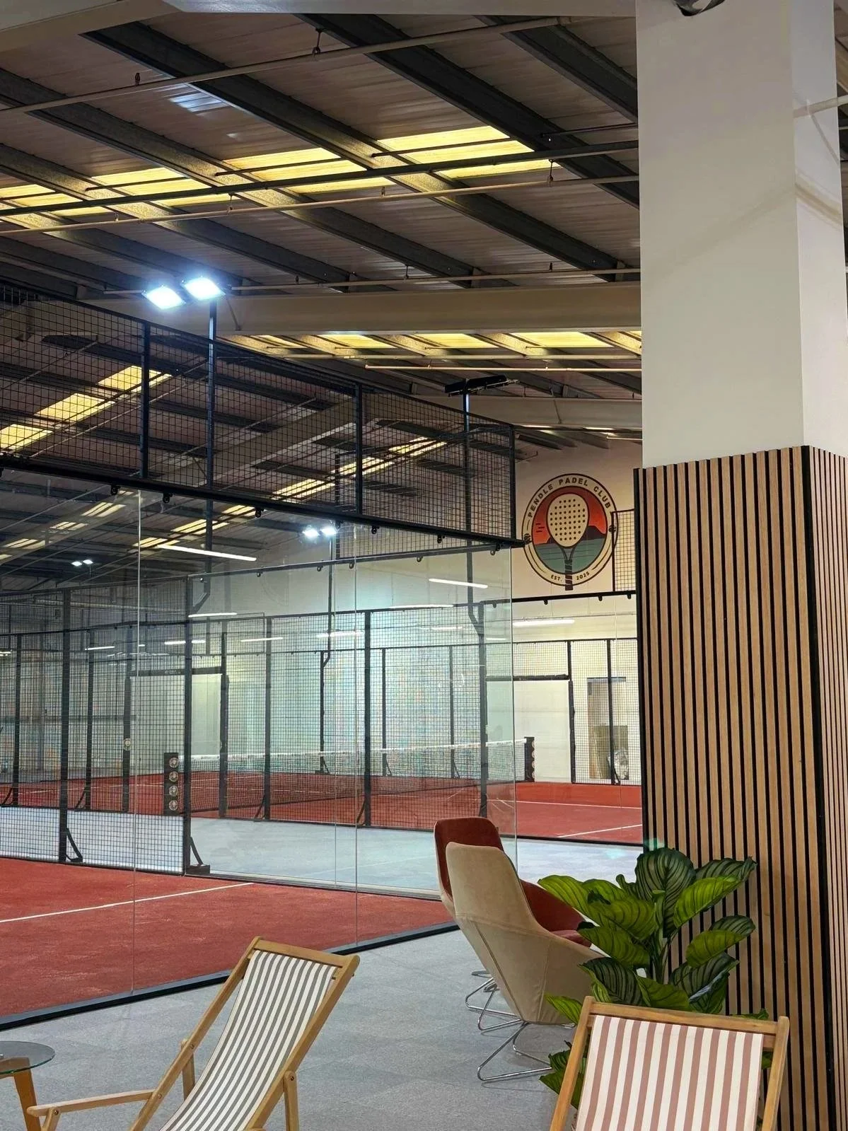 Indoor padel tennis court surrounded by glass walls, with seating area including a tan chair, a beige and a brown chair, a green plant, and wooden paneling on the right. A sign of the Padel Club with a padel and ball logo is on the wall.
