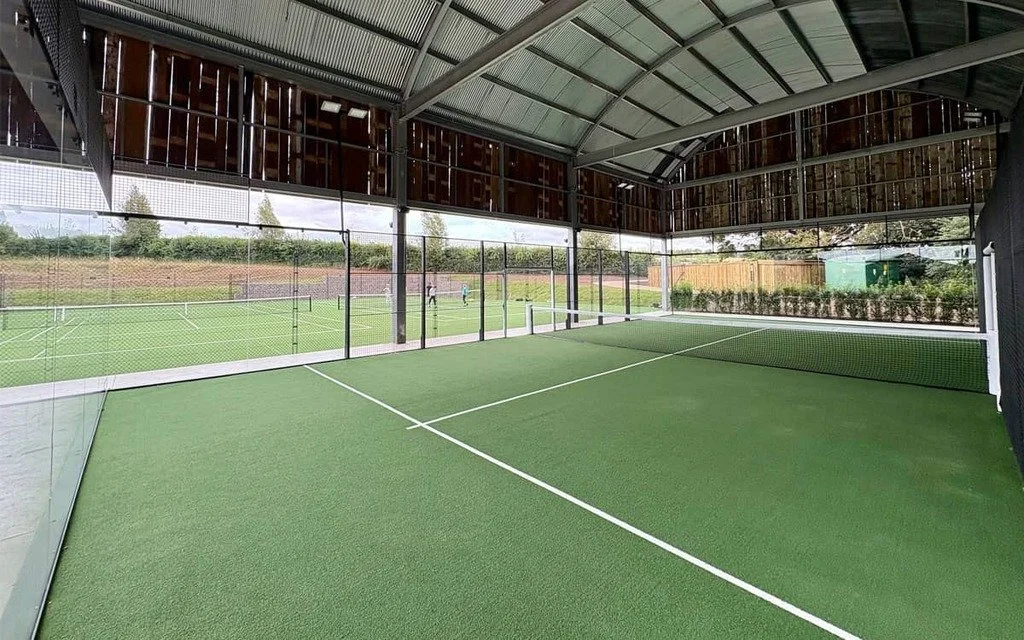 Indoor padel court with net, green surface, and large windows showing outdoor tennis courts and people playing. Soho Farmhouse