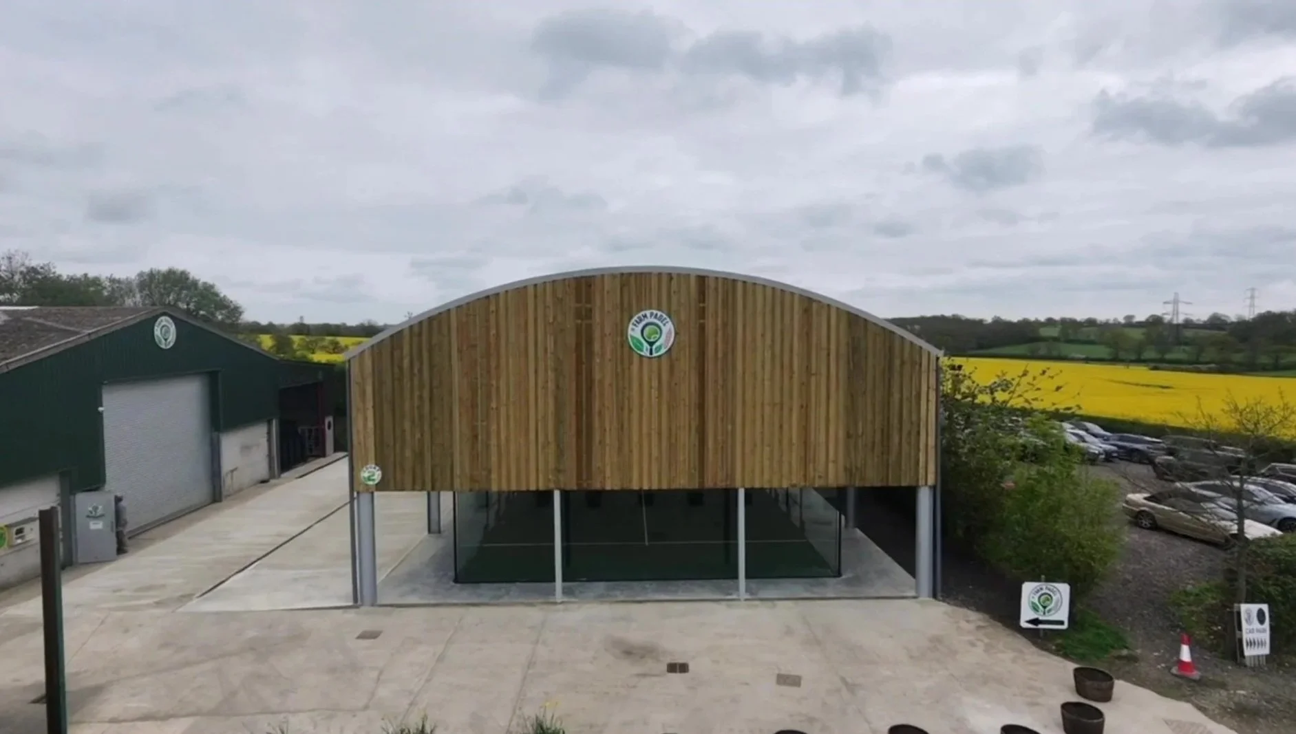 A large wooden and metal Canopy with a sign reading 'Farm Padel' with a Padel Court Underneath. This is situated in a rural area with fields of yellow flowers and greenery in the background, and a parking lot with several cars.
