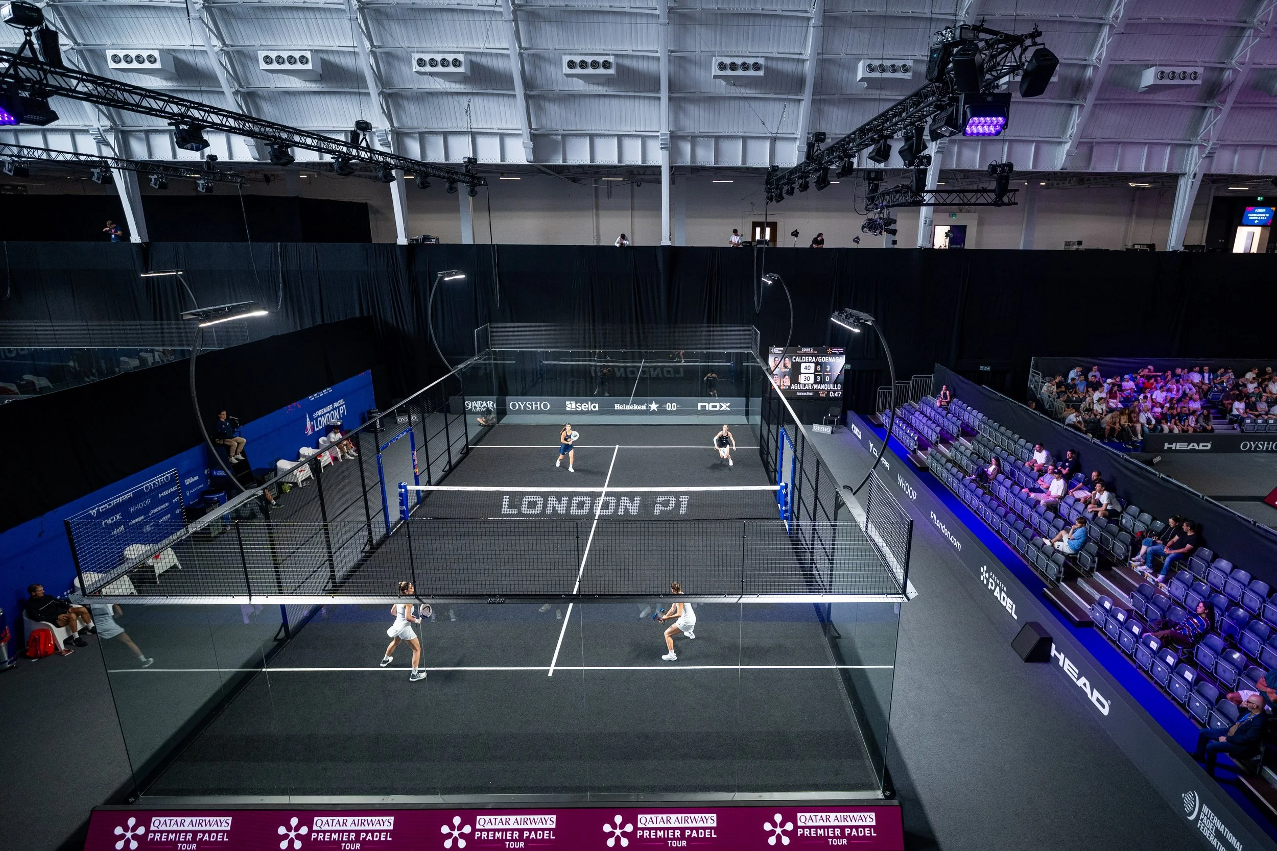 An indoor padel tennis tournament with players on the court and spectators watching from the stands. The court is surrounded by black high walls and fencing, with a digital scoreboard and advertising banners visible.
