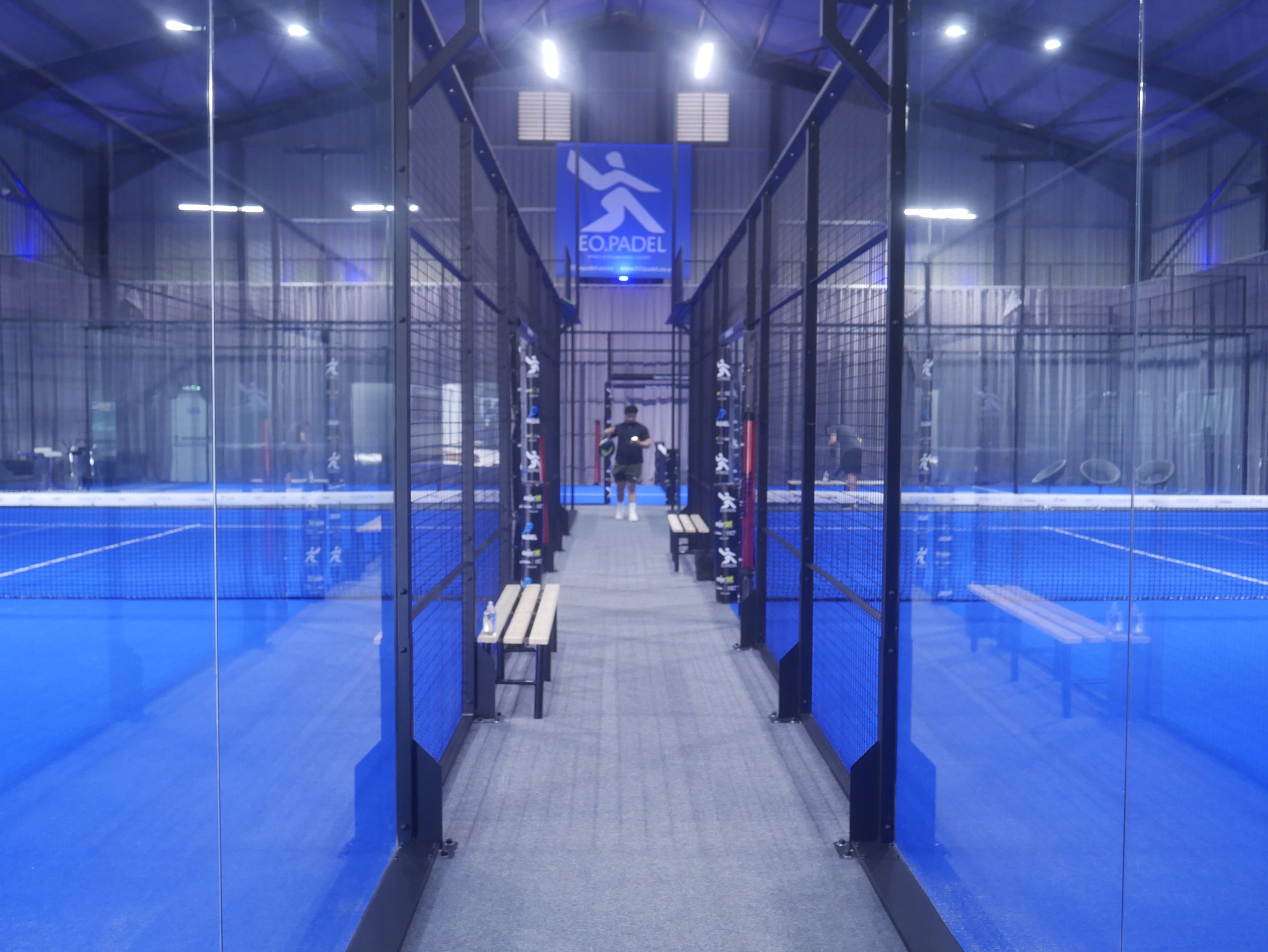 An indoor padel court enclosed by glass panels with blue playing surface. Two people are playing padel, seen through the glass. There is a blue sign with a padel icon and the text 'EO.PADEL' hanging from the ceiling.