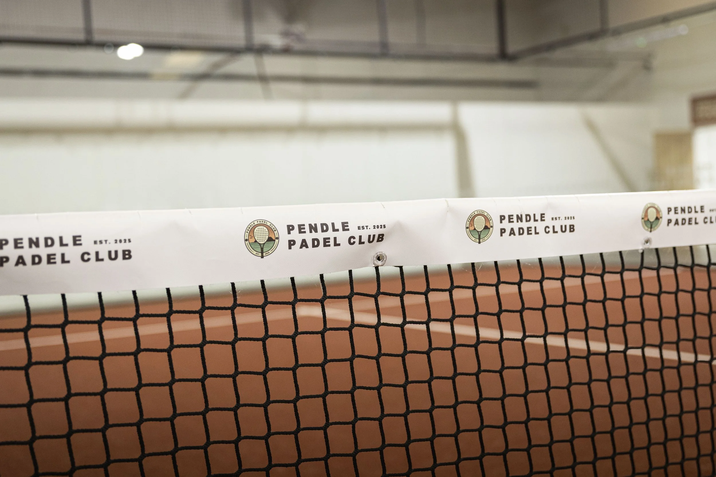A close-up view of a Padel net with a white band that has the logo and name of the Pendle Padel Club, with a blurred indoor sports court in the background.
