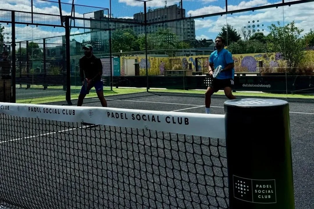 Two people playing padel tennis on an outdoor court, enclosed by glass walls, with a sign that reads 'Padel Social Club'. MejorSet