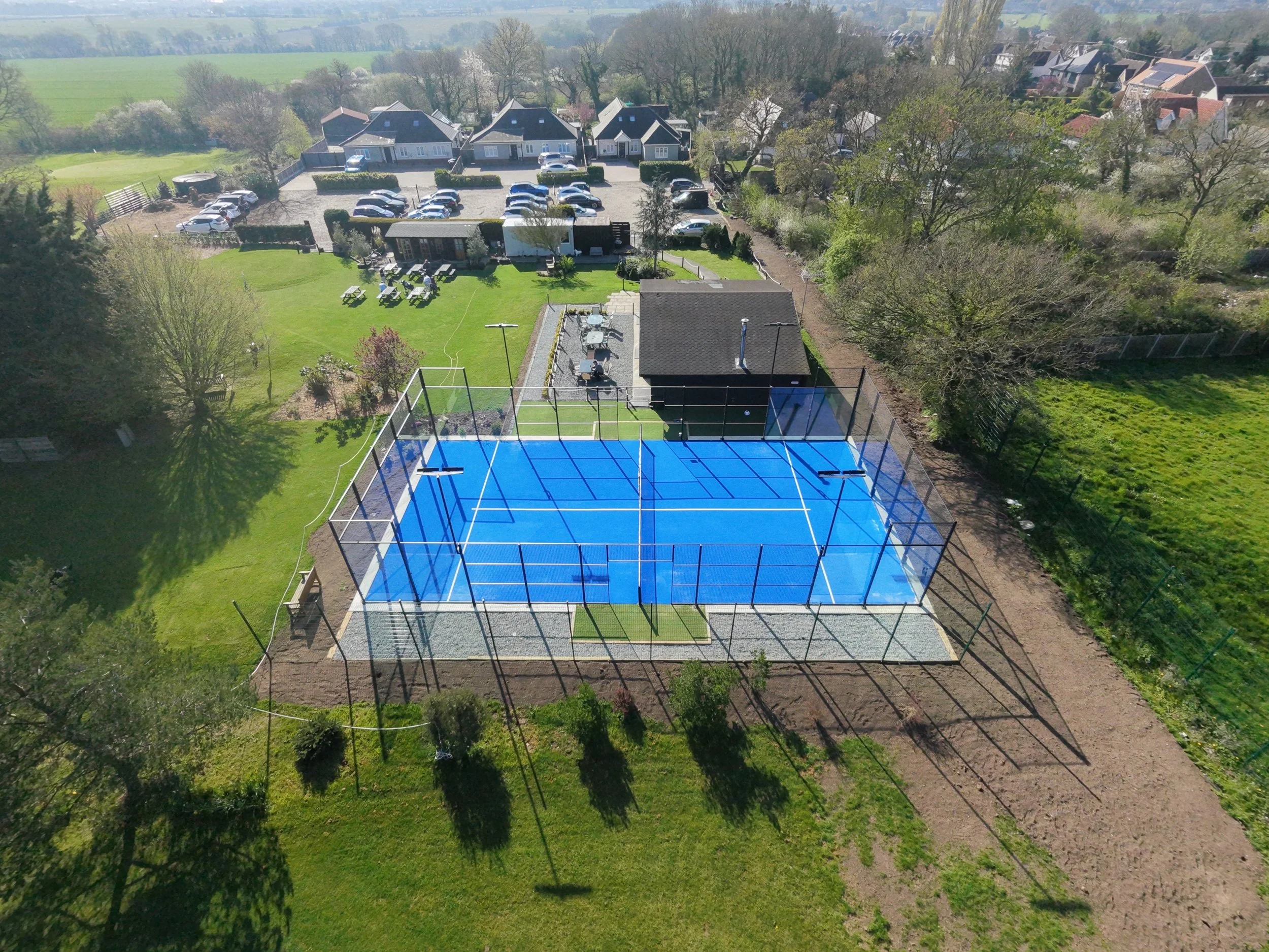 An aerial view of a residential backyard with a Padel court, a well-maintained grassy area with picnic tables, trees, and nearby houses and open fields.