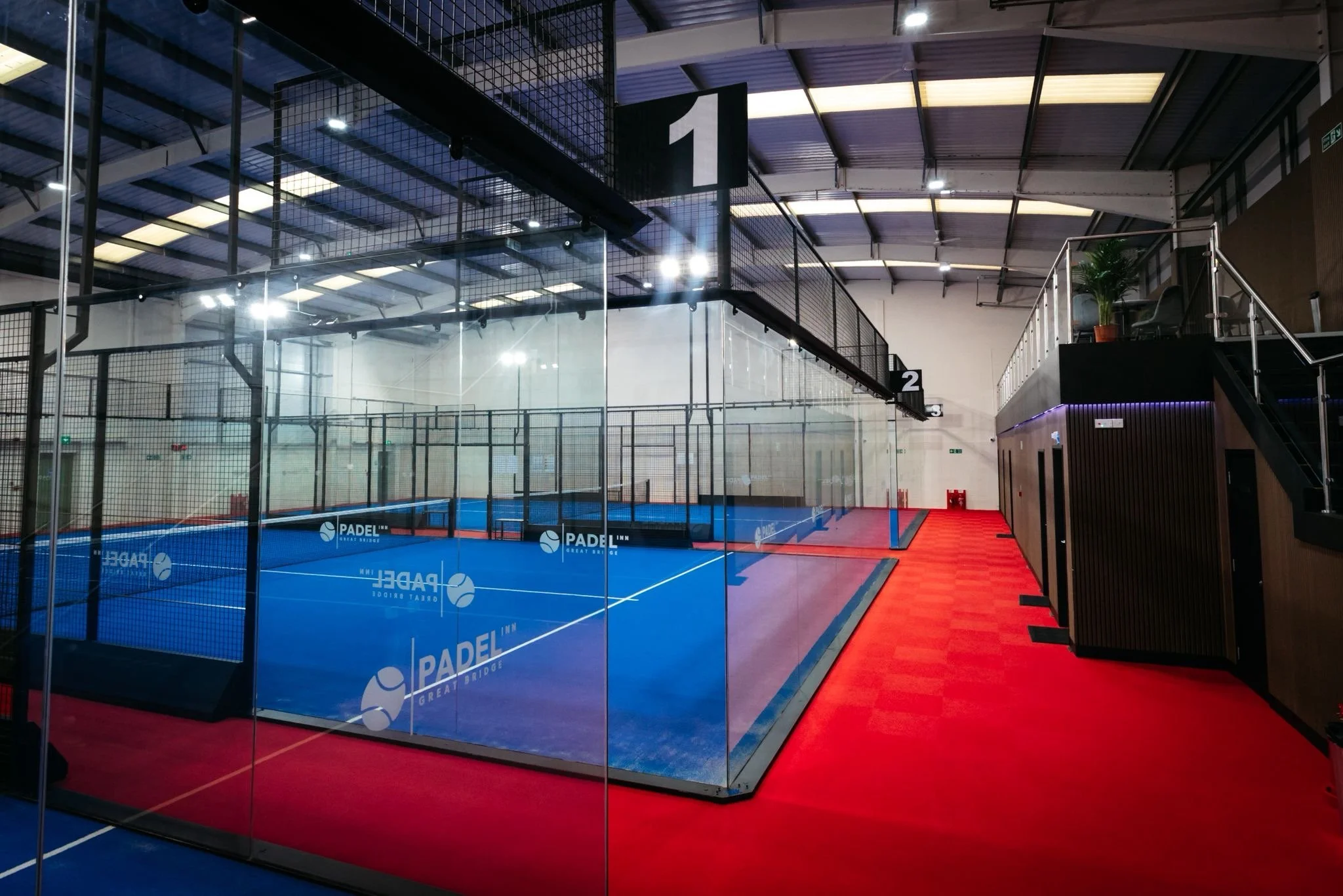 Indoor padel court with blue playing surface and glass walls, located in a modern sports facility with numbered signs, red flooring on the side.