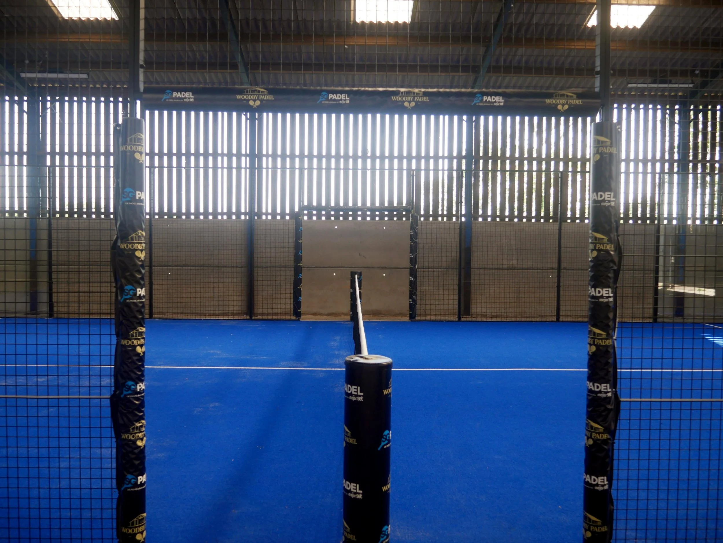 Indoor padel tennis court with blue flooring, black posts, and fence, sunlight coming through window blinds in the background.
