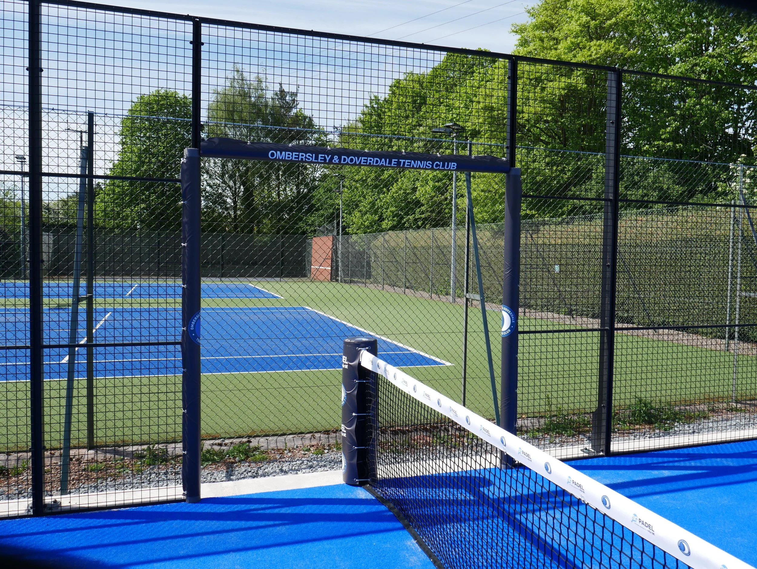Outdoor tennis and padel court at Ombersley & Doverdale Tennis Club, enclosed by a black metal fence with a blue playing surface and a white padel tennis net.