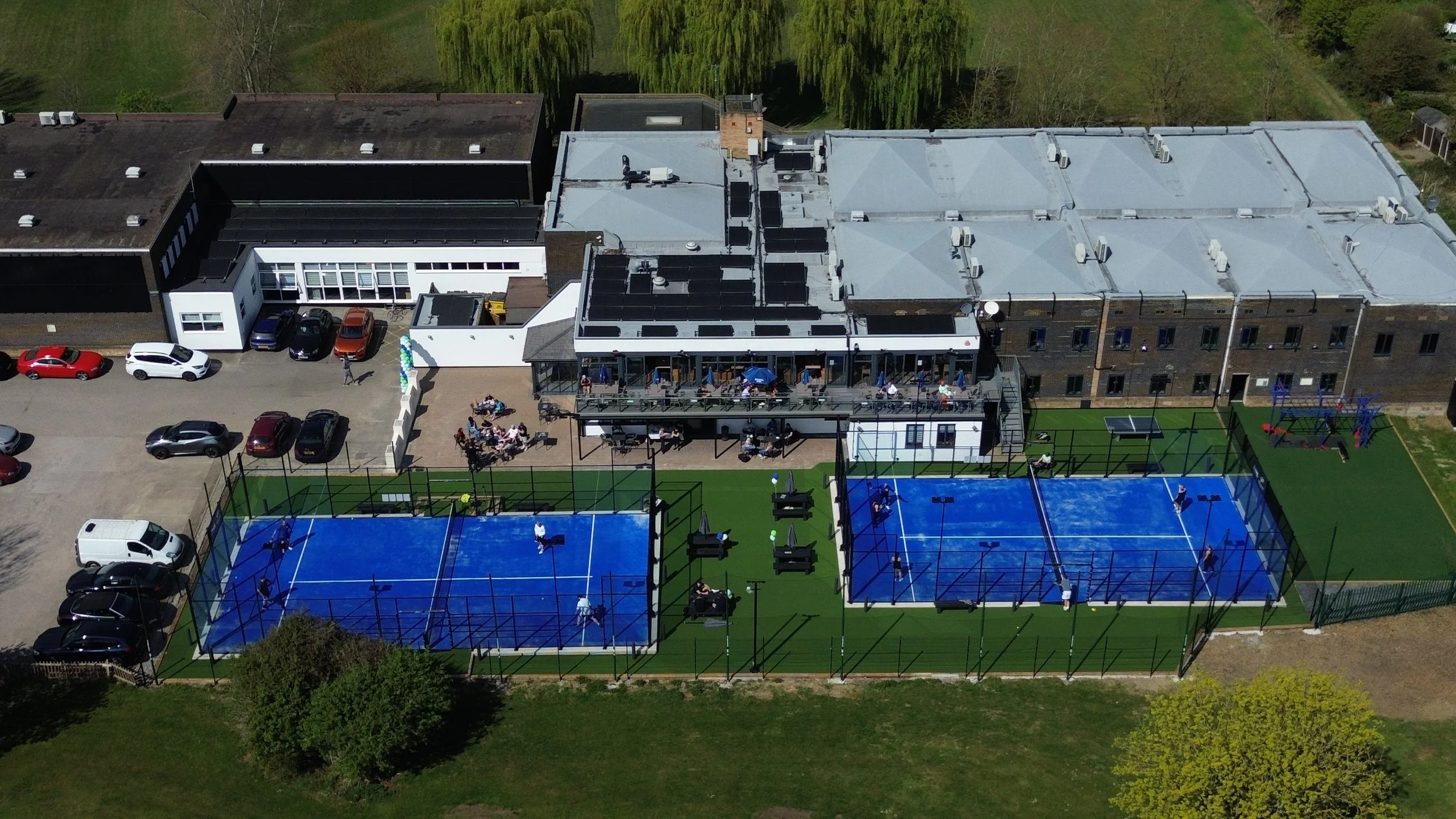 Aerial view of a recreational area with two blue Padel courts, a playground, and an outdoor seating area with people, situated next to a building with a parking lot, green trees, and a grassy field.