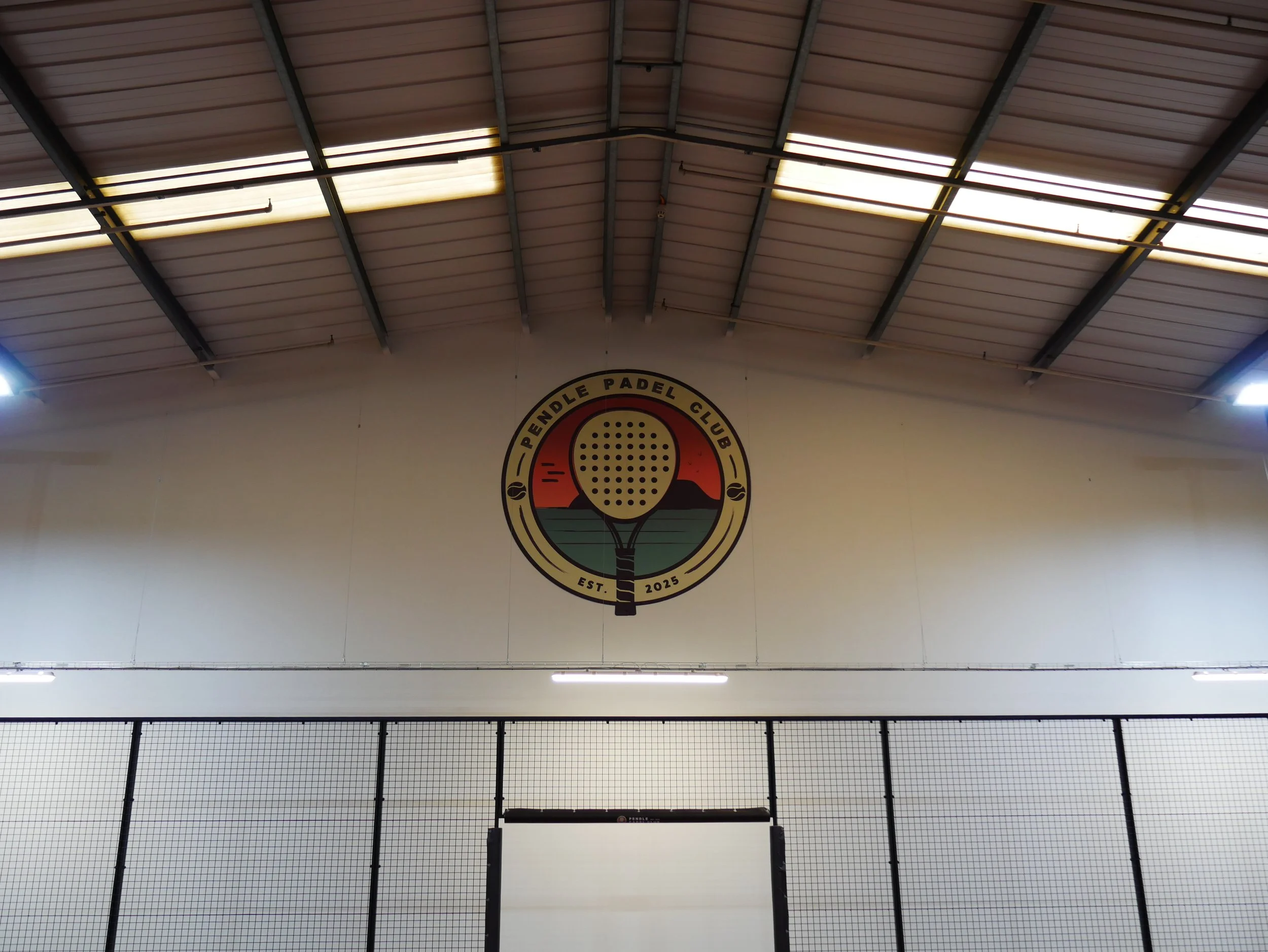 Indoor Padel facility ceiling with metal beams, translucent panels, and a wall with a circular logo for a padel club.