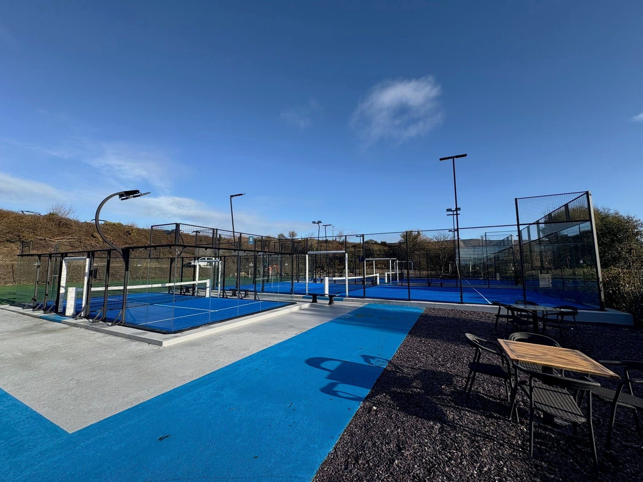 Outdoor sports courts, including pickleball and tennis, with tables and chairs nearby, under a clear blue sky with a few clouds.