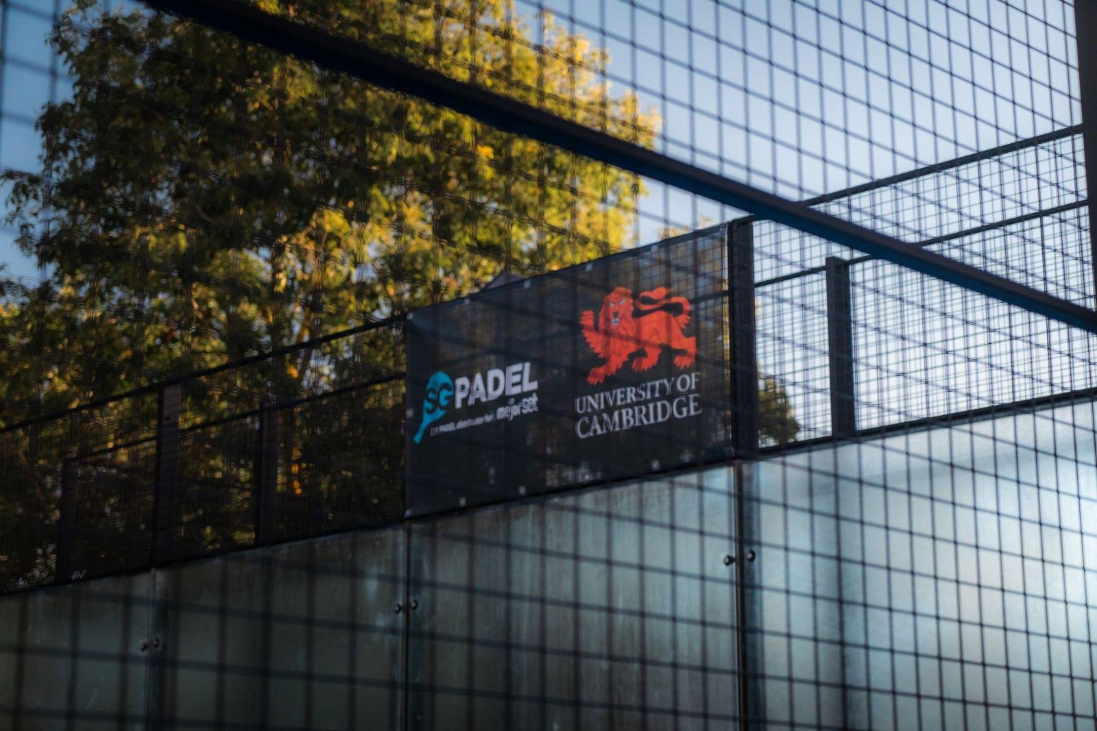 A building with a glass exterior reflecting trees and blue sky. There are banners displaying the logos of SG Padel and the University of Cambridge.
