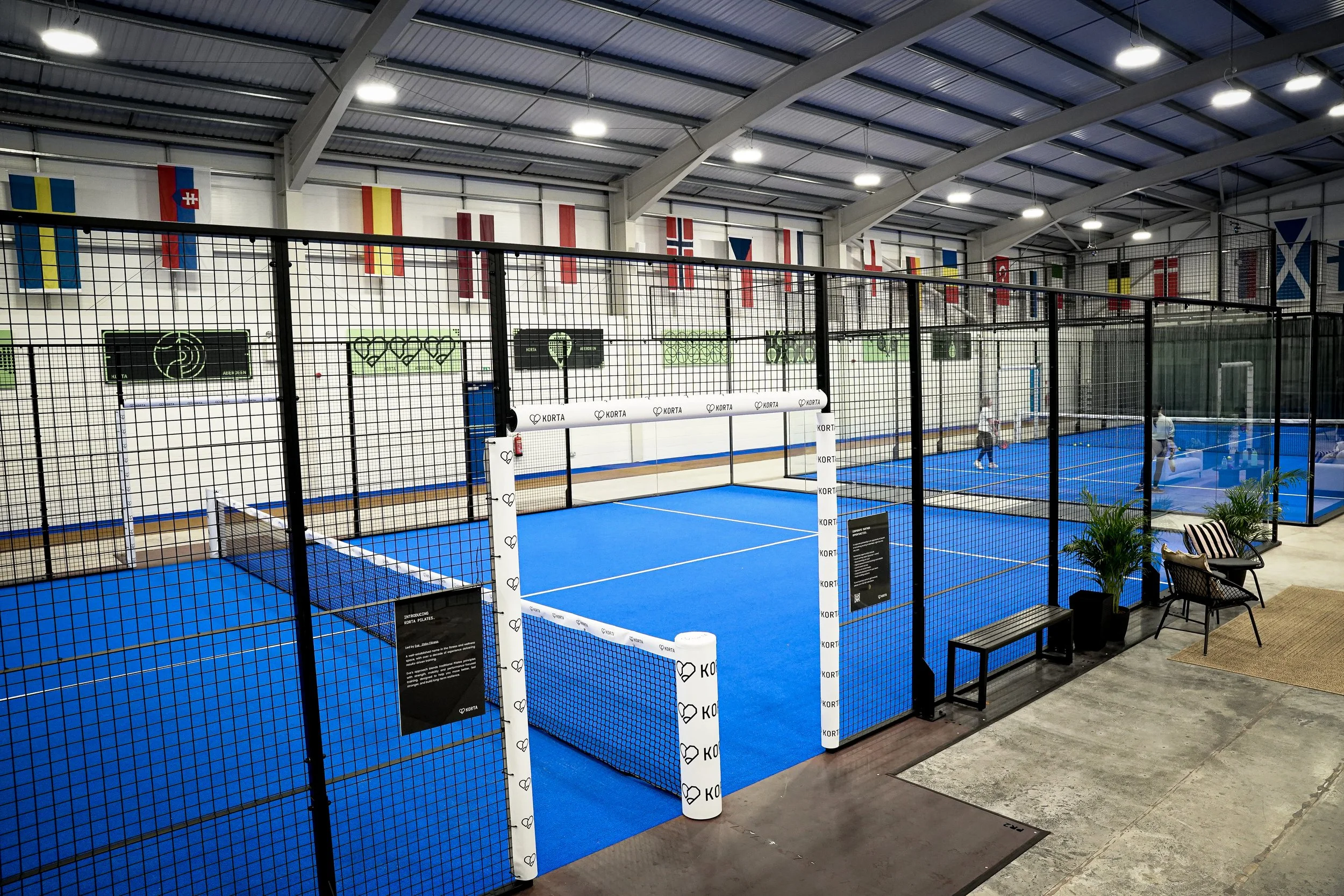 Indoor sports facility with blue courts enclosed by black netting, flags from various countries hanging from the ceiling, and players practicing on the courts. 4 Pop up Padel Courts