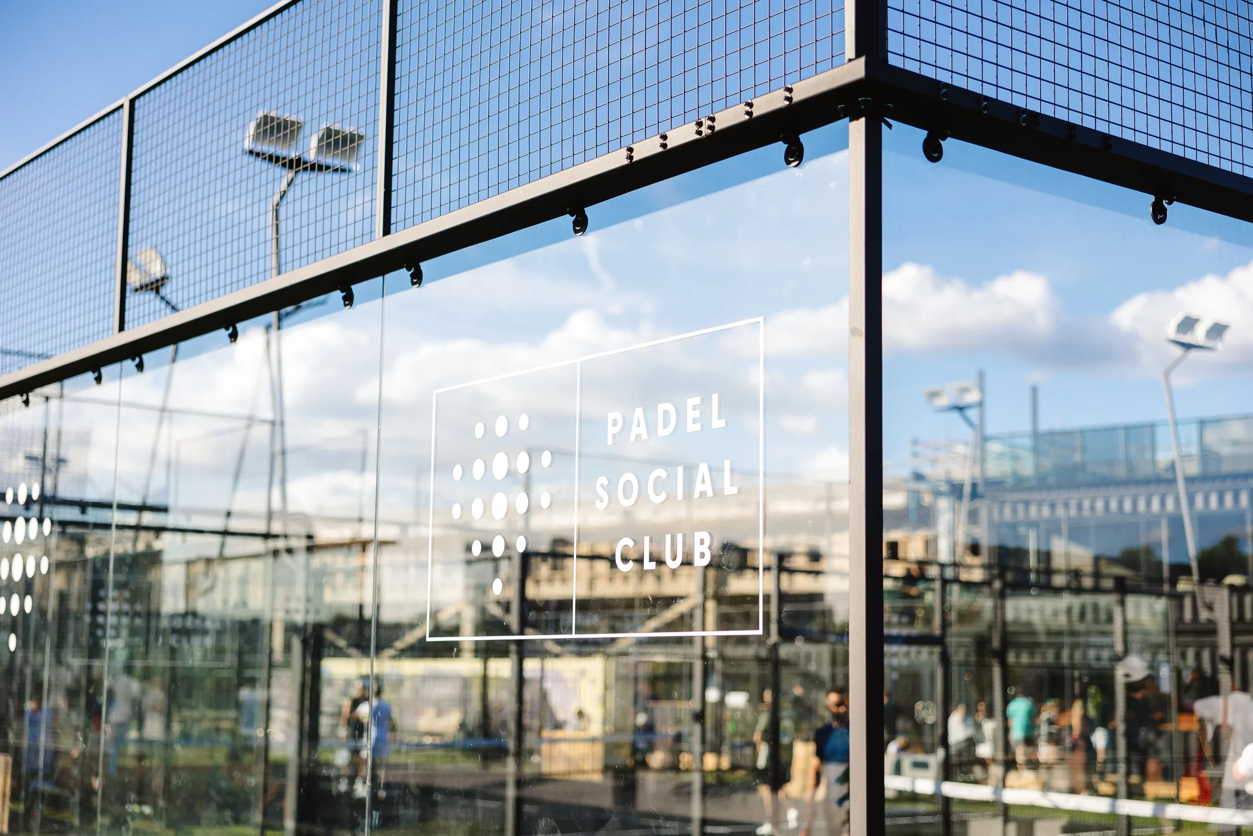 Glass enclosure with the words 'Padel Social Club' printed on it, with outdoor sports court and people in the background under a partly cloudy sky. UK Padel