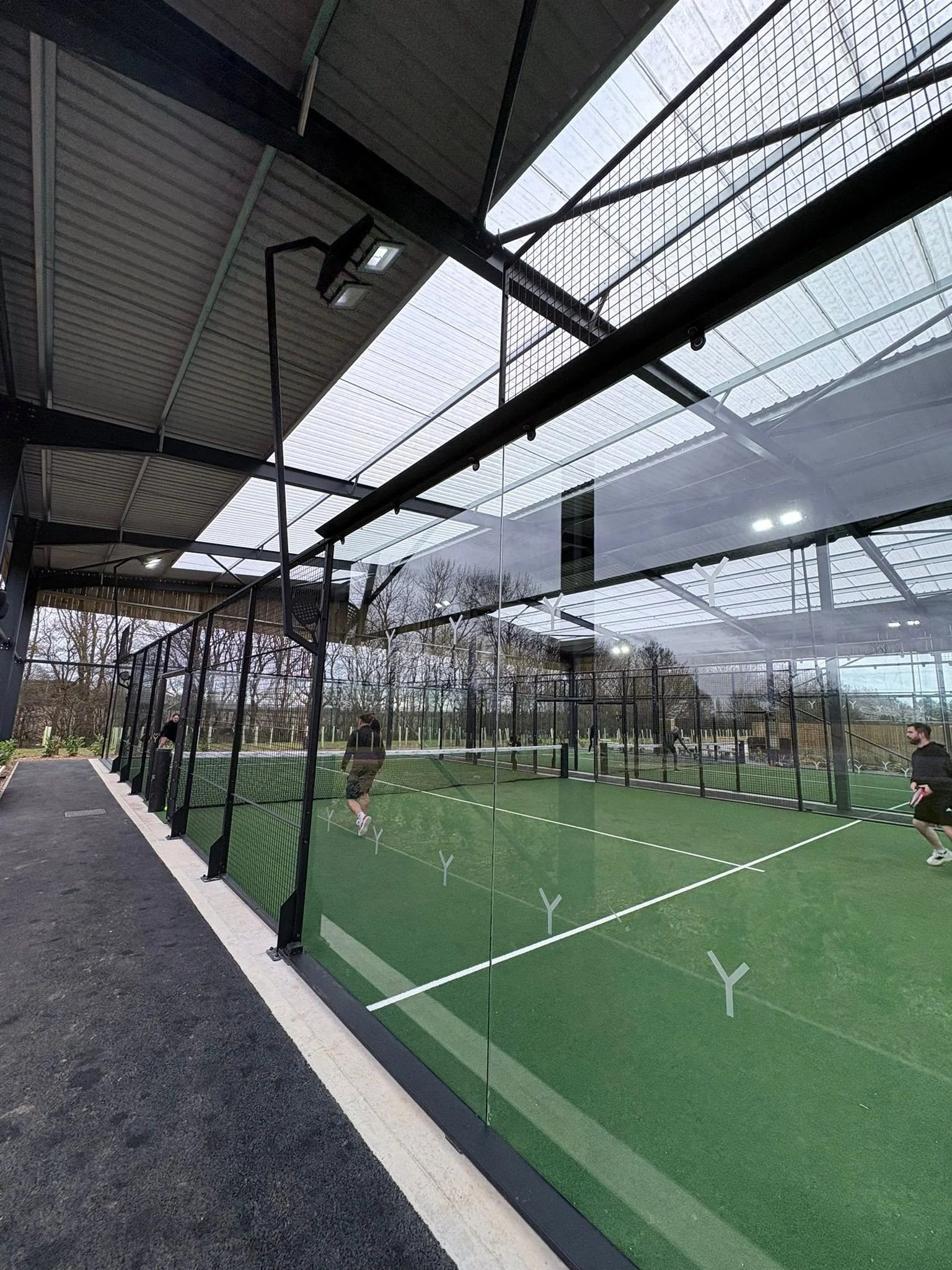 Indoor Padel court with a green artificial turf surface, surrounded by glass walls, and a roof with skylights. Several people are playing Padel, and there are trees visible outside.
