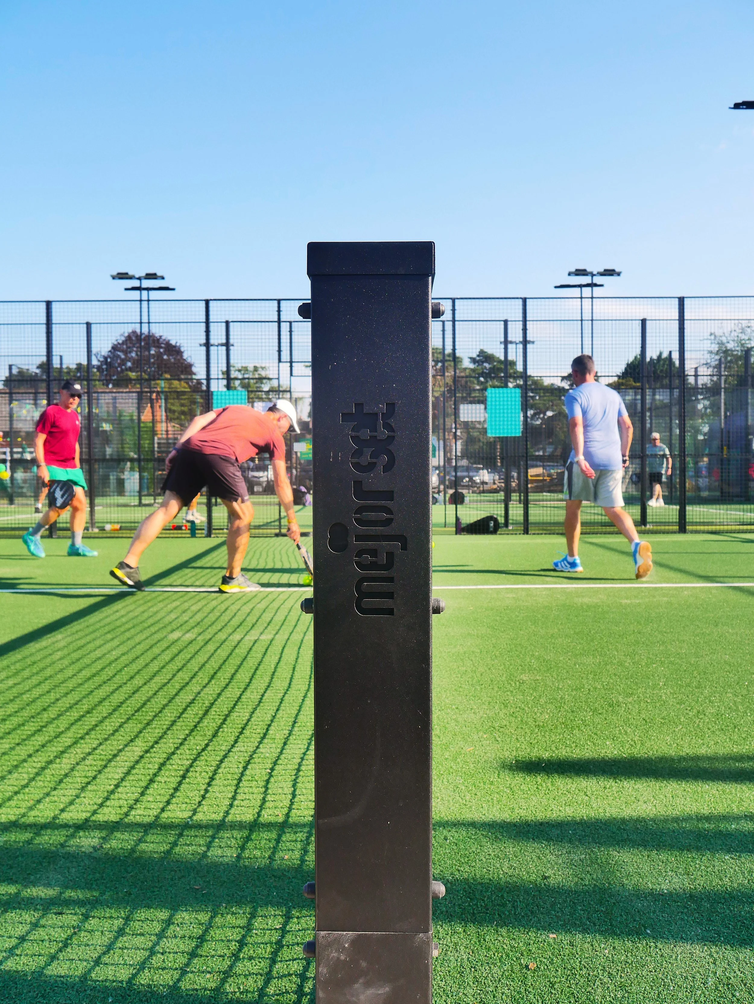 An outdoor pickleball court with a black net post in the foreground, players playing in the background, and bright sunlight casting shadows on the court.