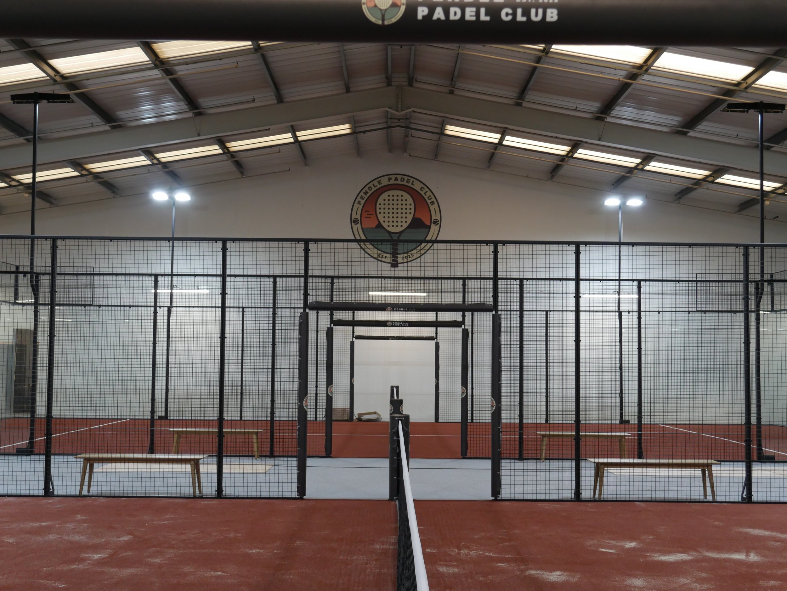 Indoor padel tennis court with Red Terracotta with a black padel racket logo at the top. The court is enclosed with black wire fencing, and there is a white padel racket positioned at the center of the court, with chairs on either side.