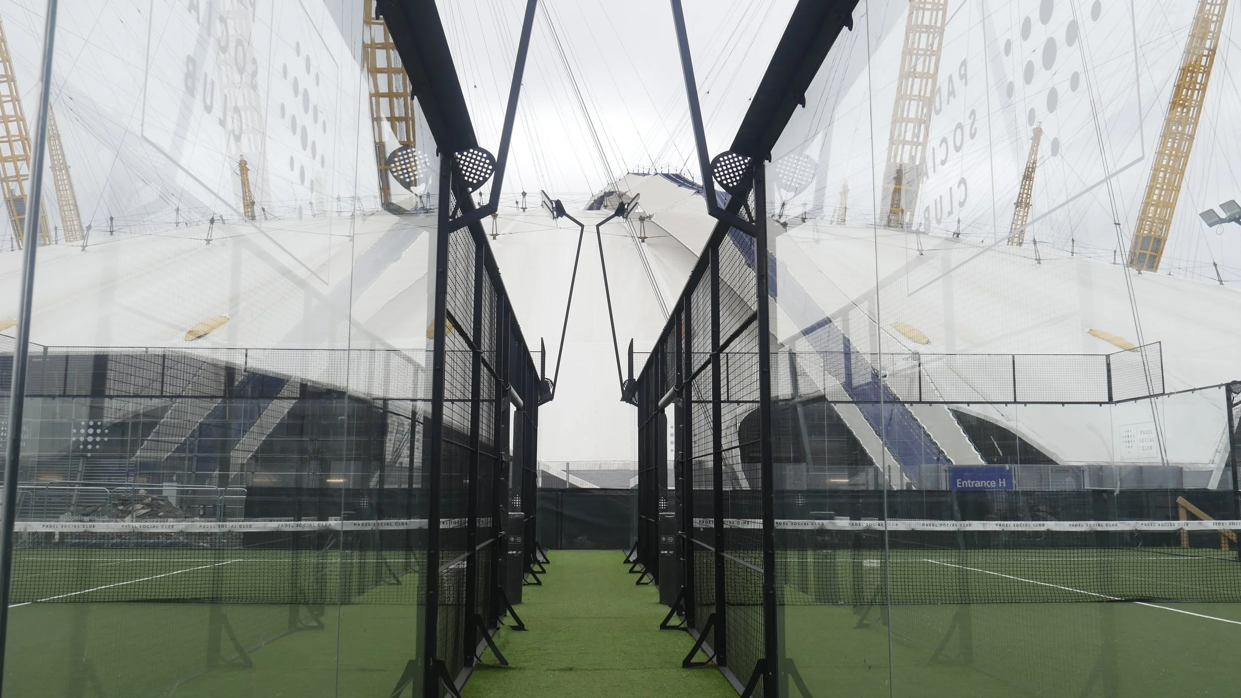 An indoor padel tennis court with a glass wall and black fencing. The court has artificial green turf, and the structure is visible through the glass. There are black barriers and signs, and the overall setting appears modern and enclosed.