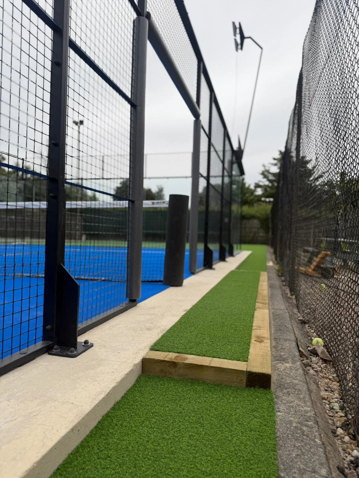 A close-up shot of a fenced tennis court with a blue surface, surrounded by black chain-link fences. There is a narrow strip of artificial green grass alongside the concrete sidewalk.