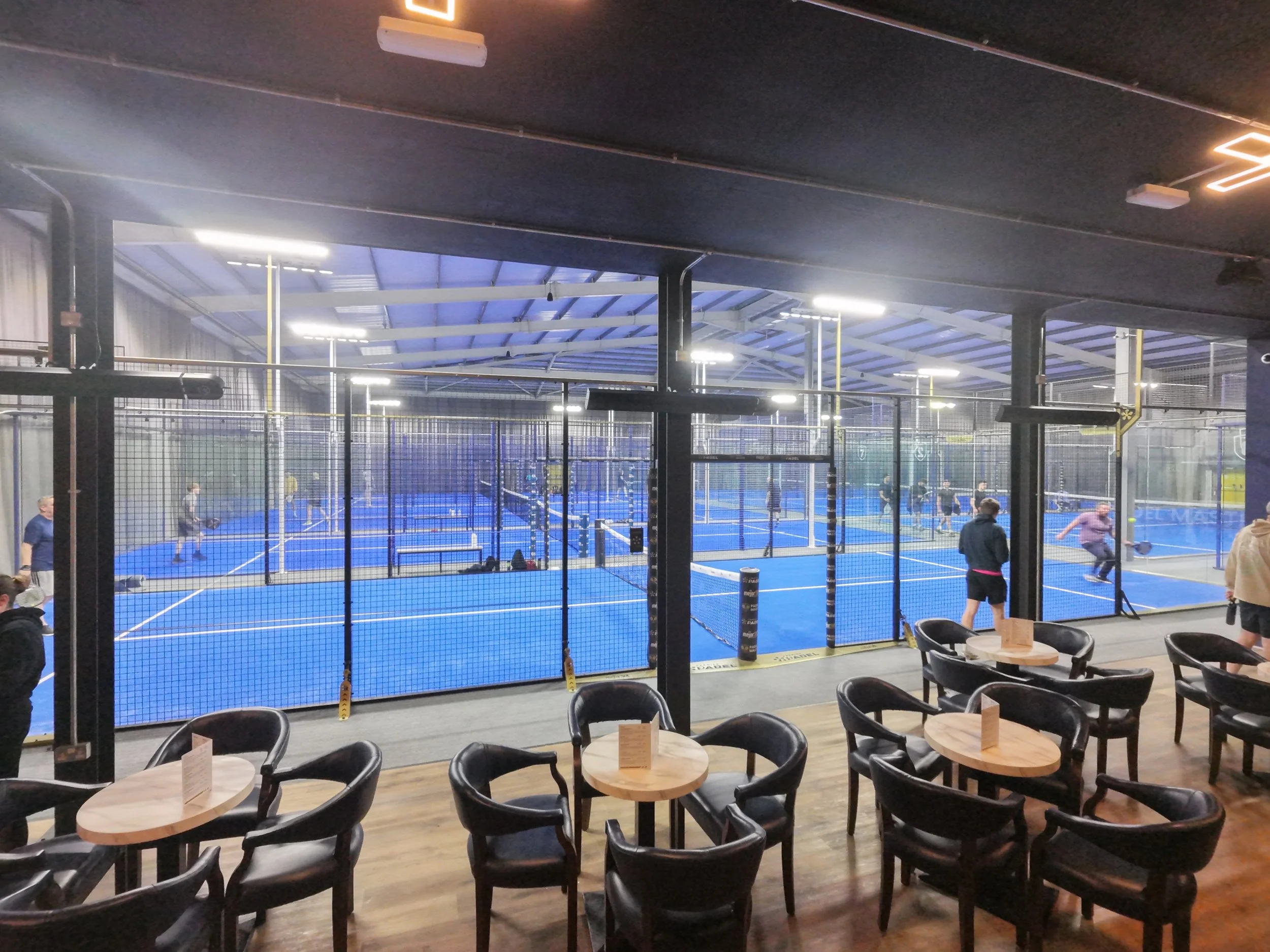 Indoor sports facility with blue Padel courts, surrounded by a black fence, and people playing. In the foreground, there are wooden tables with black chairs around them, inside a lounge or café area with some people seated.