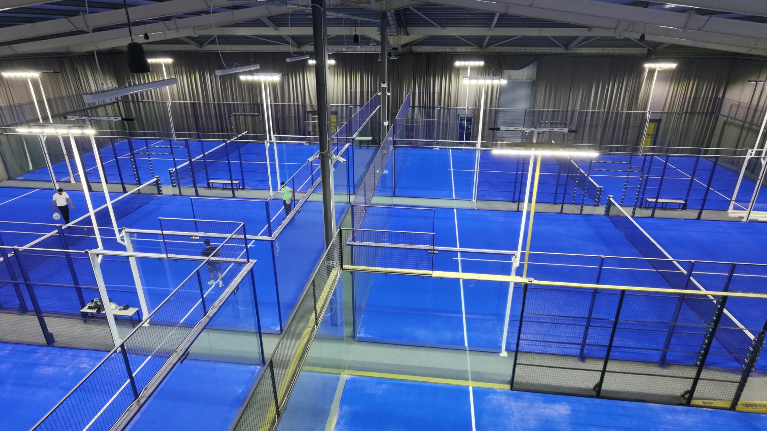 Indoor sports complex with multiple blue padel tennis courts enclosed by black fences, with two players practicing on one court, surrounded by bright overhead lights.