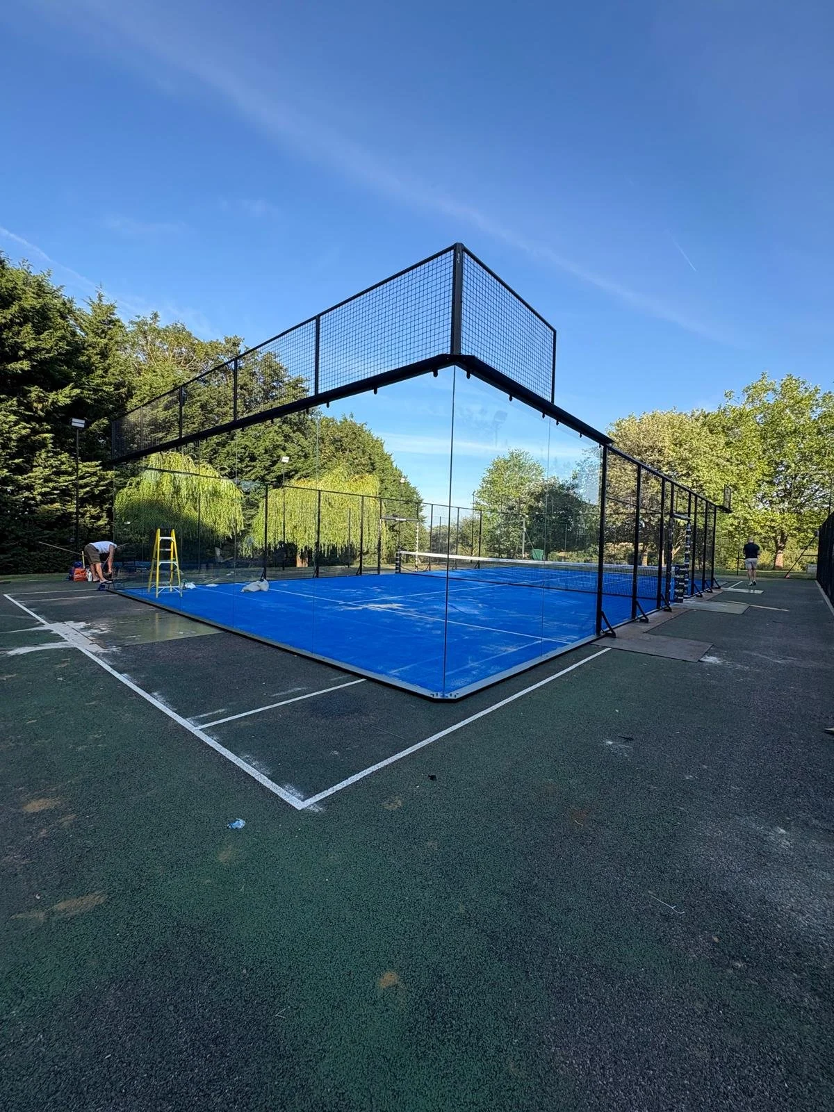 A tennis court with a blue surface is under construction, surrounded by black fencing. Workers are installing or repairing the court, with one person on the left and another on the right. The court is situated outdoors, with trees and a clear blue sky in the background.