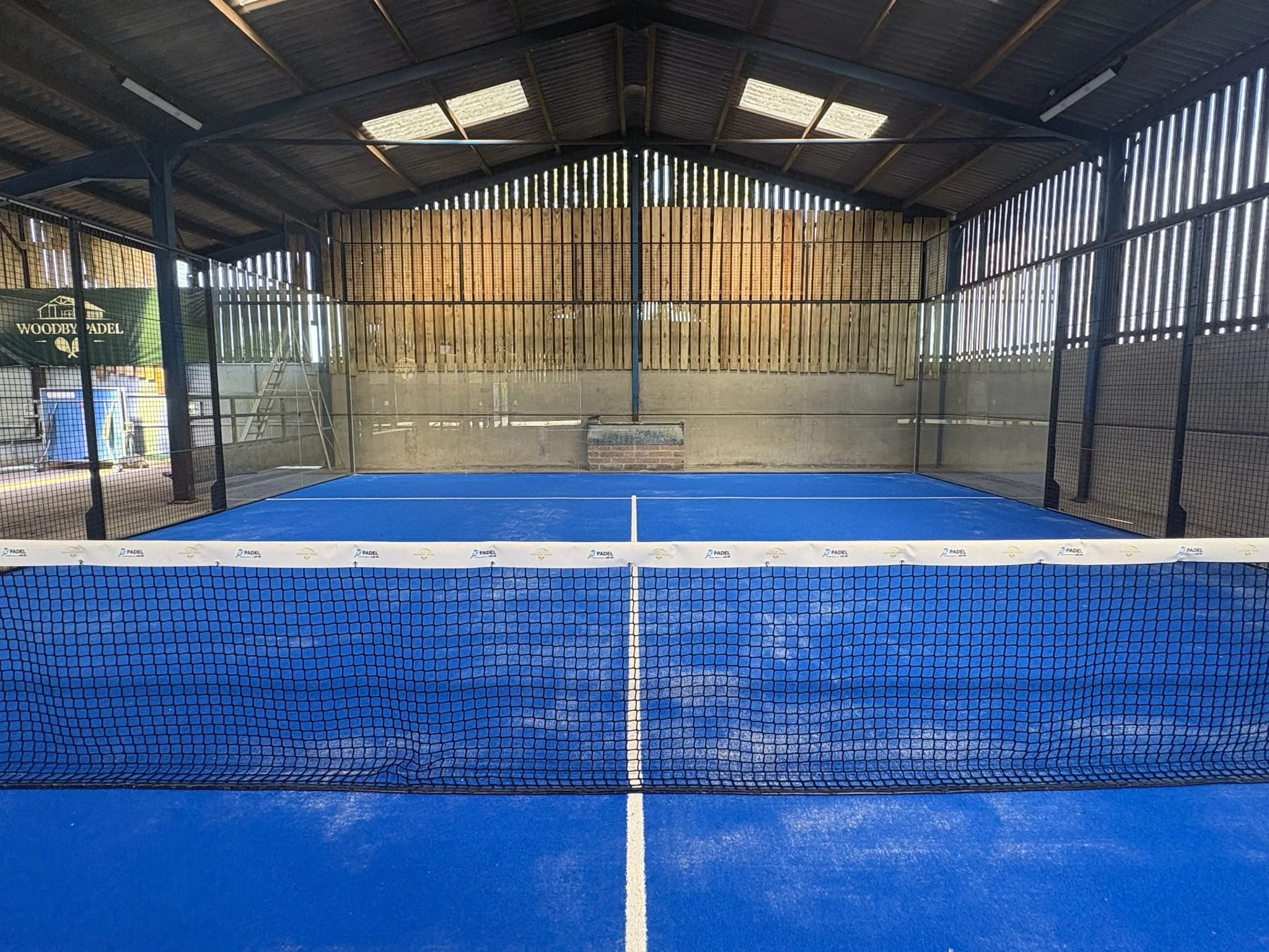 Indoor padel tennis court with blue surface, black protective fencing, and a wooden wall at the back. The roof is made of metal panels with skylights.