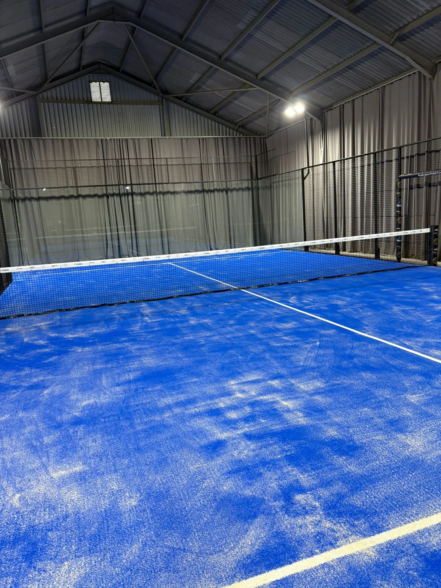 Indoor padel court with blue surface, net in the center, and curtains along the back wall.