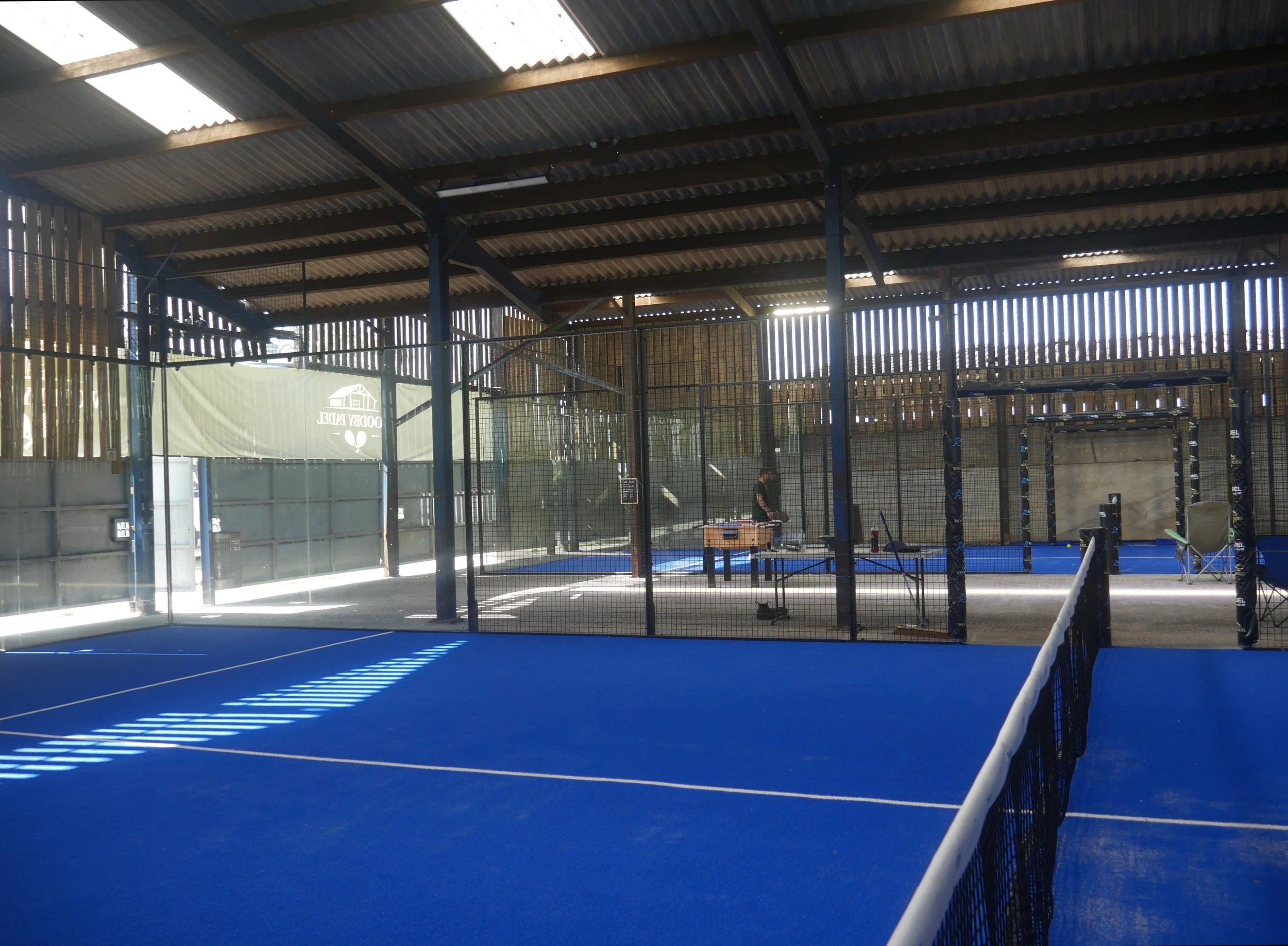 Indoor tennis court with blue surface, net, and protective fencing, sunlight filtering through skylights in metal roof.