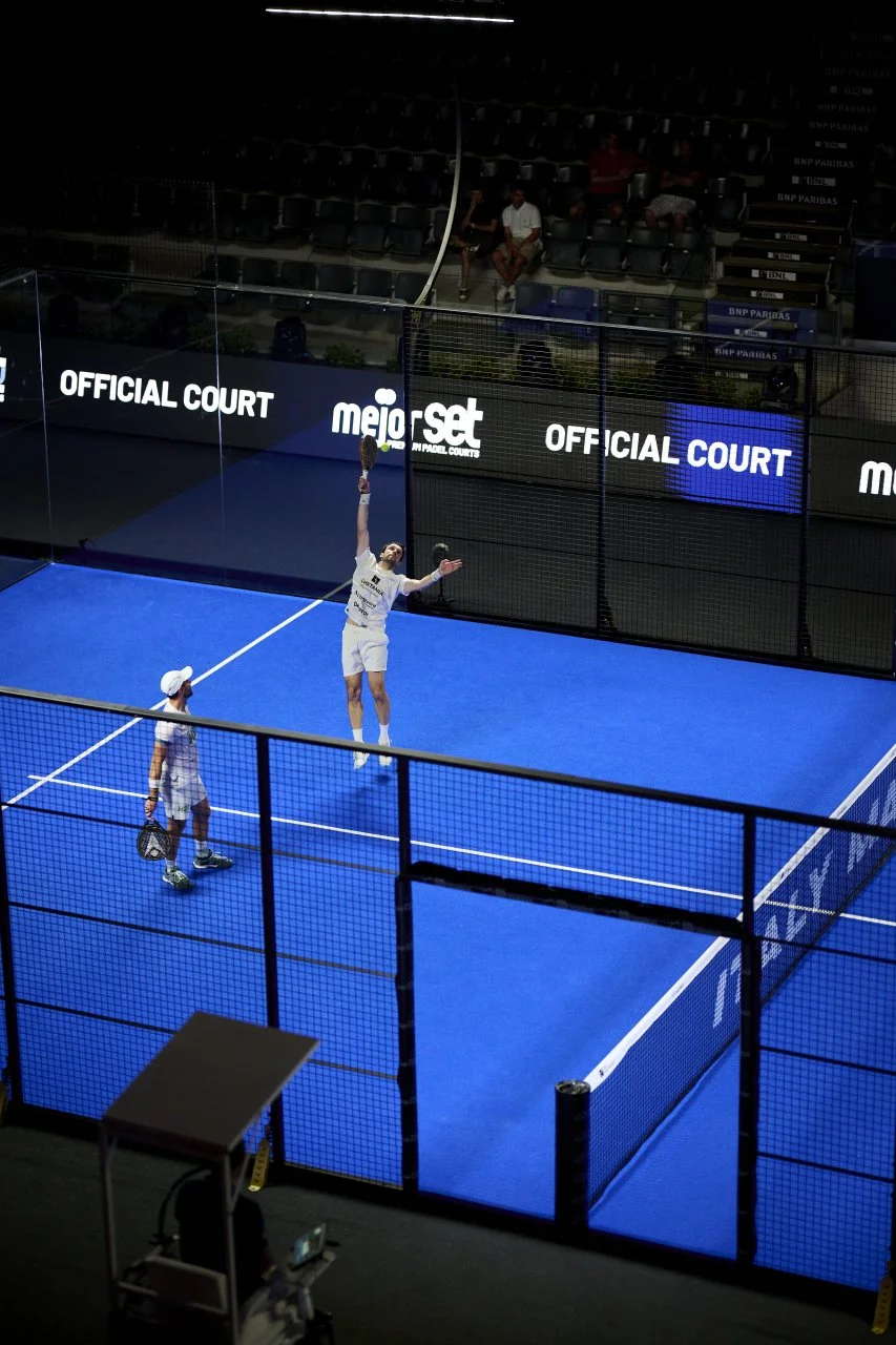A mixed doubles Padel match on an indoor court with blue surface, two players in white uniforms, one preparing for a serve. Spectators seated in the background.