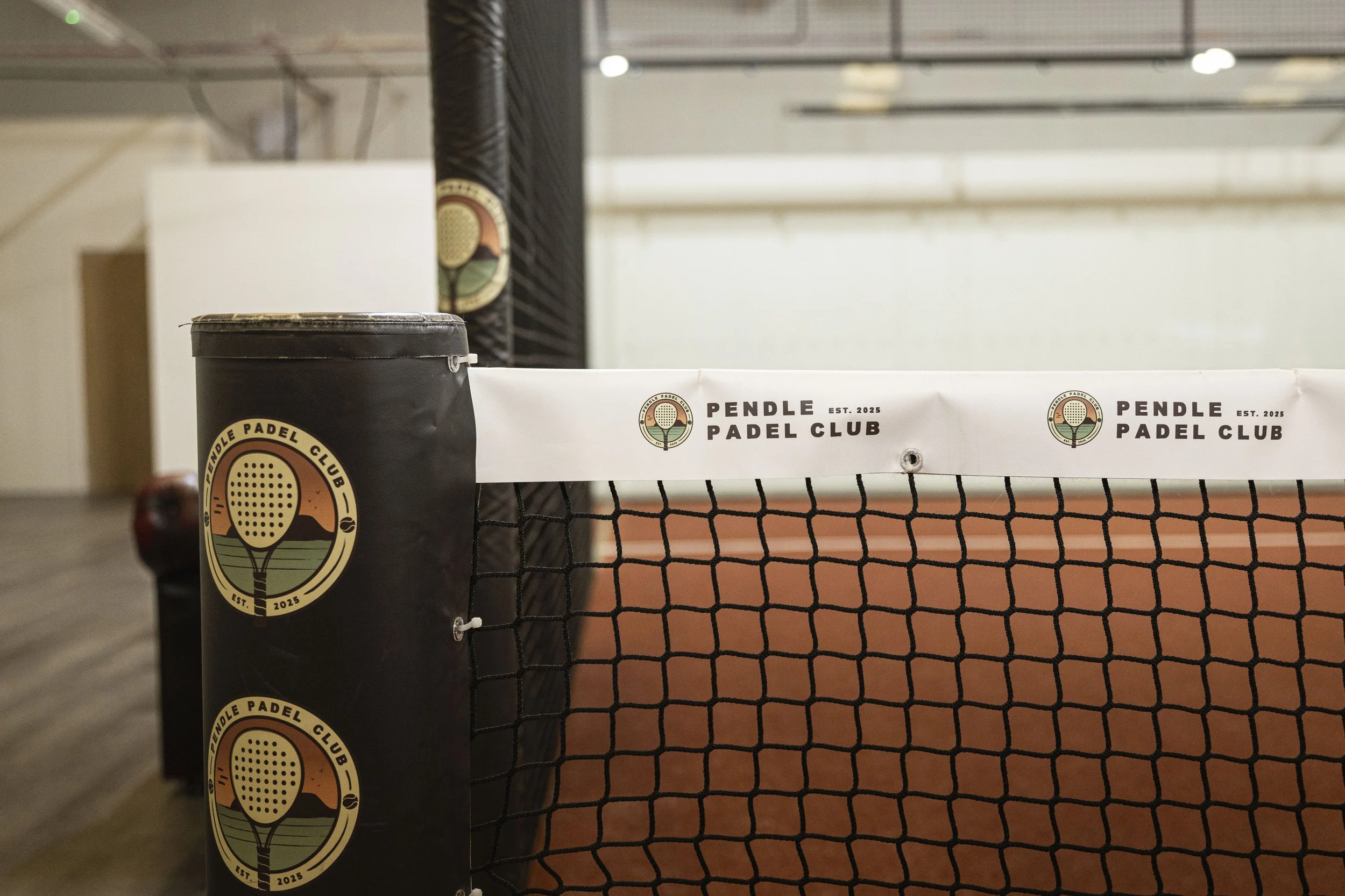 Indoor padel tennis court with a black padded post and a banner that reads 'Pendle Padel Club' with logo, net, and a blurred background of the court.