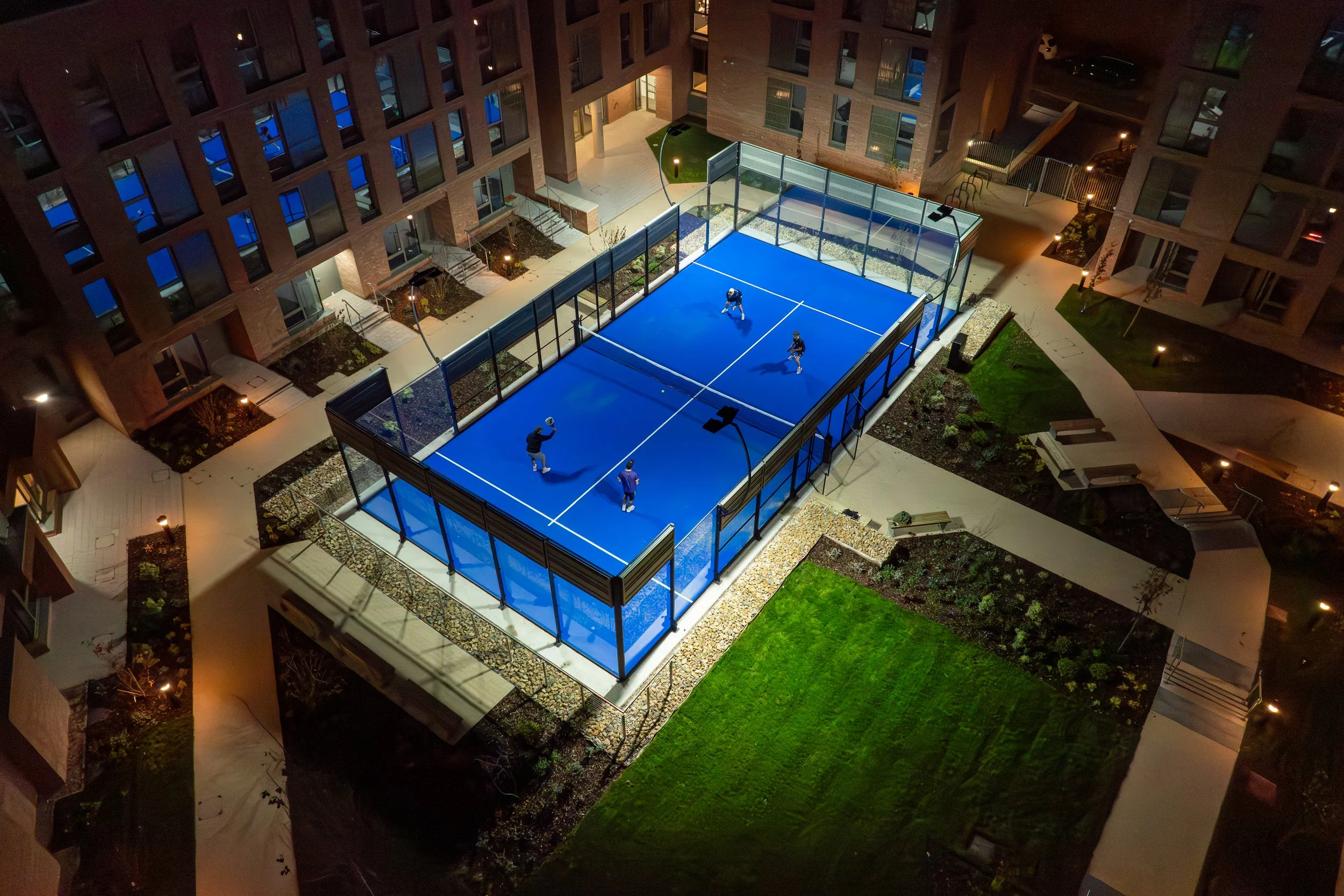 Nighttime aerial view of a rooftop tennis court with a blue playing surface, enclosed by glass walls, situated in an apartment complex courtyard with walkways, landscaped garden beds, and benches.