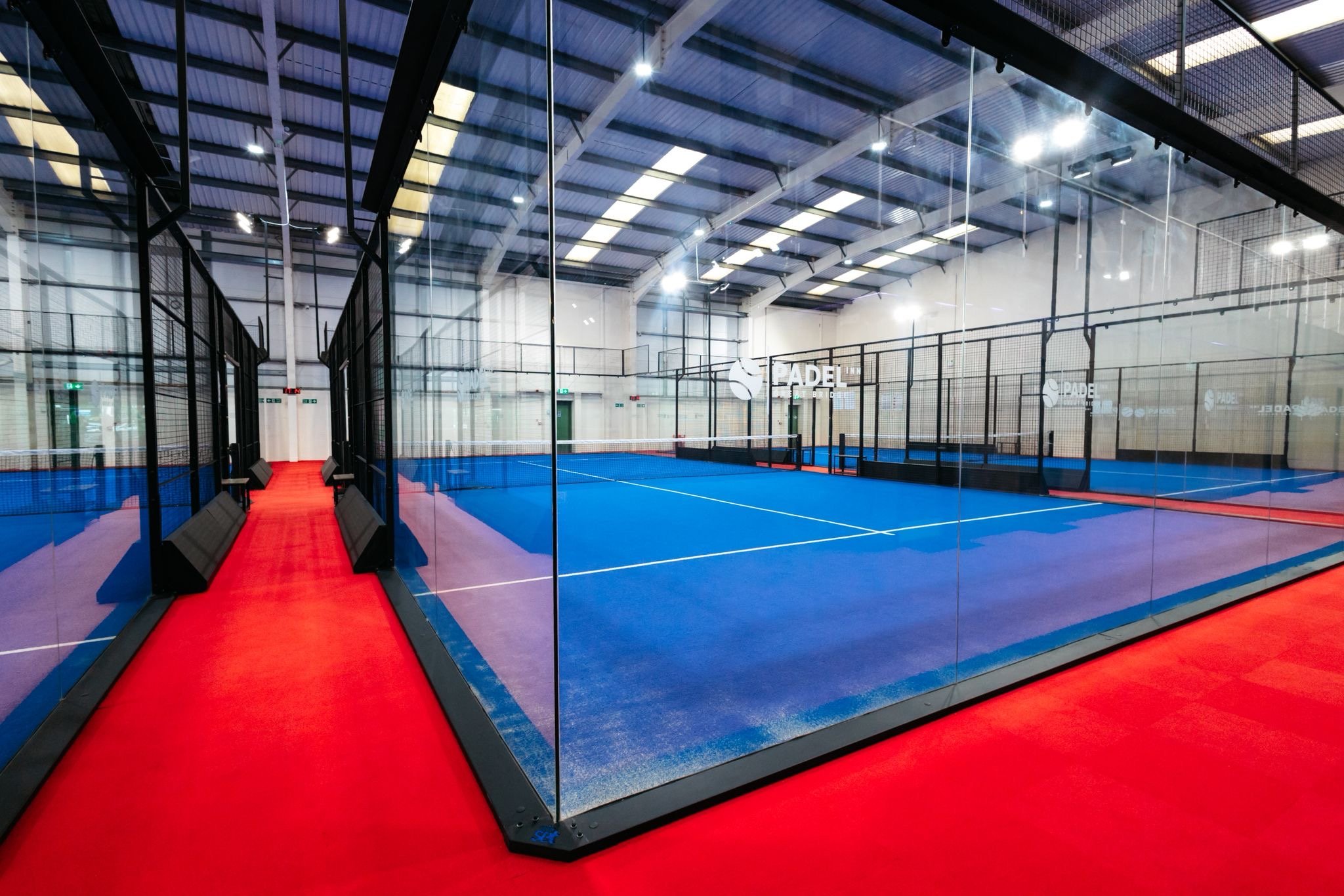 Indoor padel tennis court with blue playing surface surrounded by glass walls and black fencing UKs Best Padel