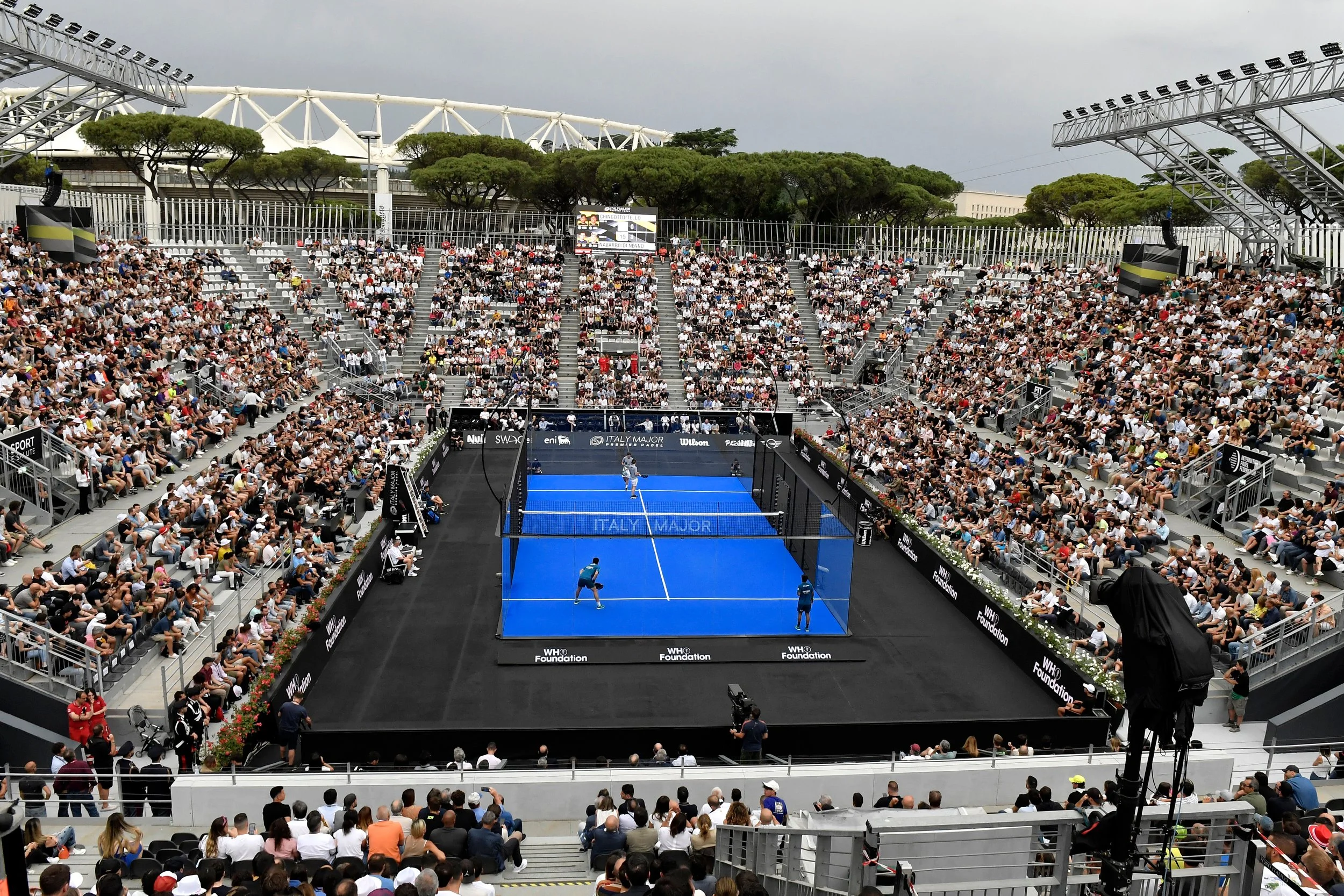 A crowded outdoor Padel stadium with spectators watching a match on a blue court. The match appears to involve doubles teams, and the venue has a modern, open design with a large roof structure and green trees in the background.
