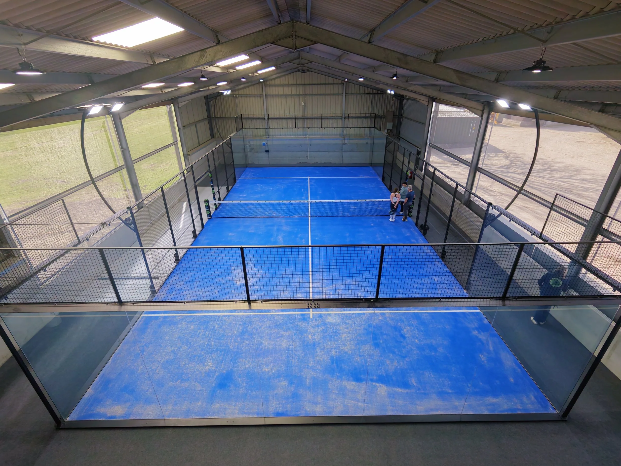 Interior of an barn with a Padel court with blue flooring, surrounded by glass walls and a metal roof. There are two people standing and talking near the corner of the court.
