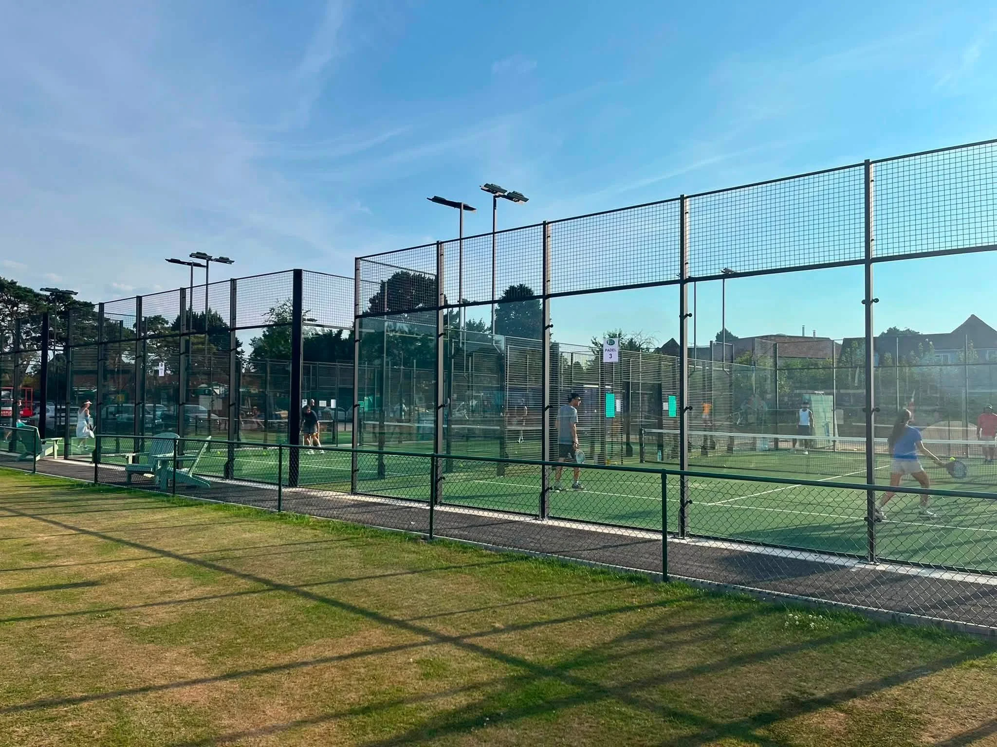 Outdoor Padel facility with multiple courts enclosed by tall fences, under a clear blue sky, with some water fountains visible in the distance.