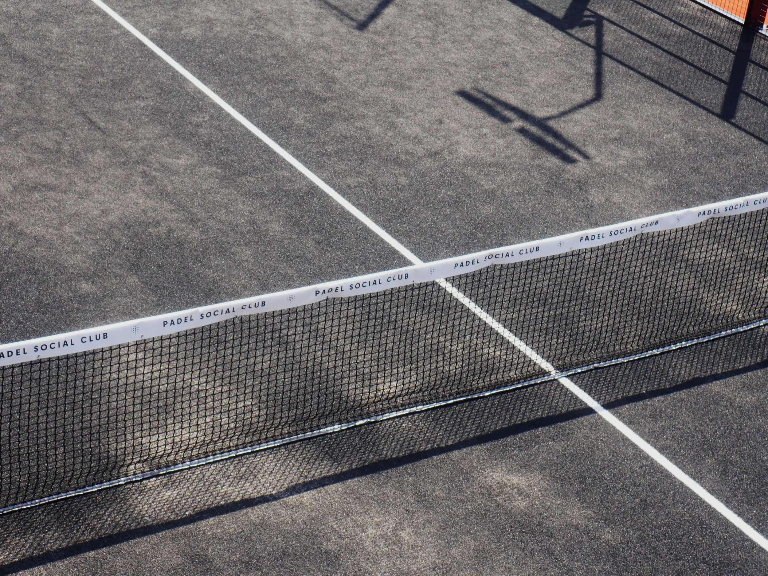 A section of a padel tennis court, showing the net with a white band that reads 'Padel Social Club' and a Black turf court surface.