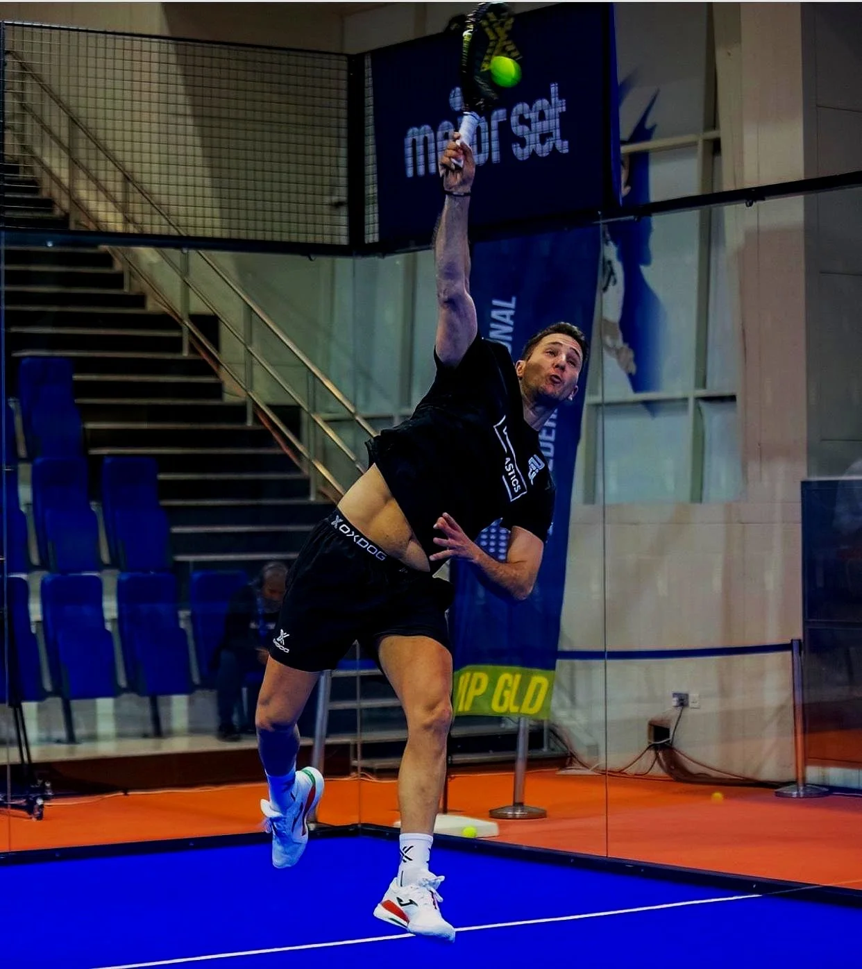A Padel Player, reaching up with a racket to hit a green ball against a glass wall, in an indoor sports facility with blue court and seating.