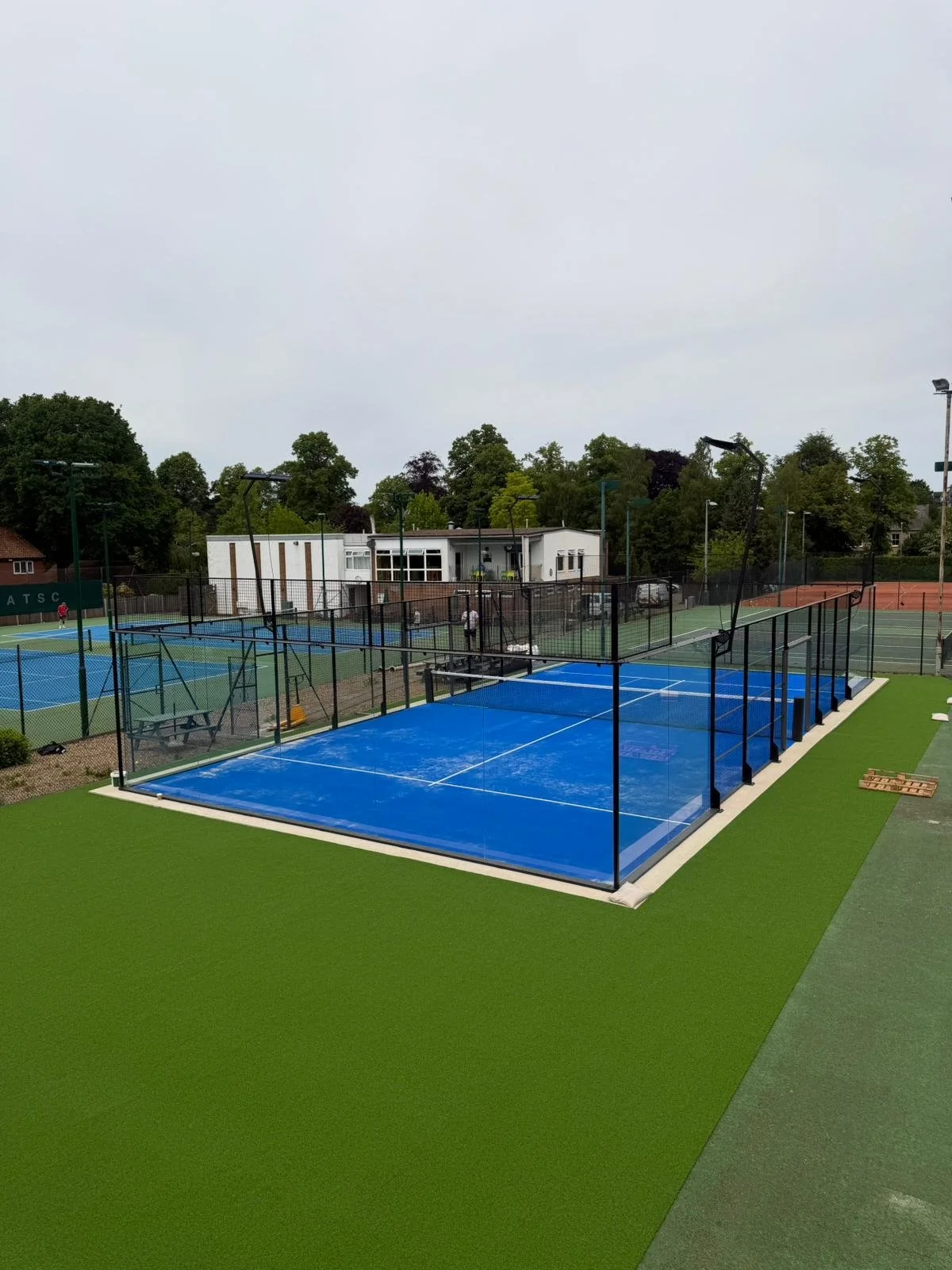 Tennis courts with green and blue surfaces, surrounded by black fencing, in an outdoor sports complex with trees in the background.