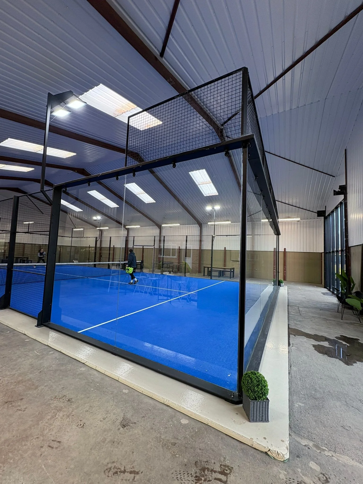 Indoor padel court with a blue surface, surrounded by glass walls, in a warehouse-like structure with a curved metal ceiling and fluorescent lights.