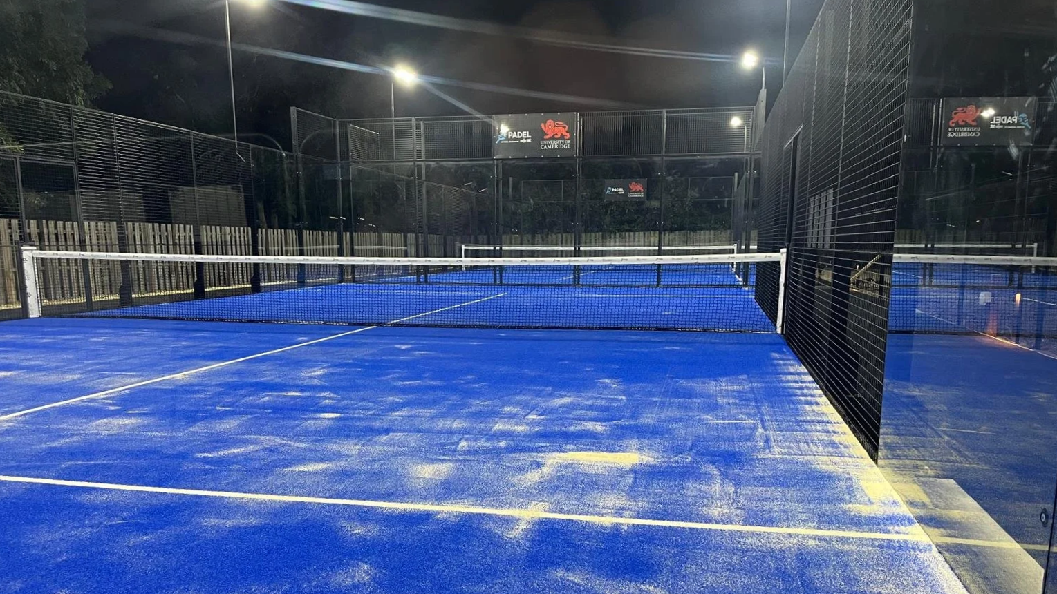 Padel Court at the University of Cambridge at night, illuminated by the court lights. Blue turf. UKs Best Padel Courts