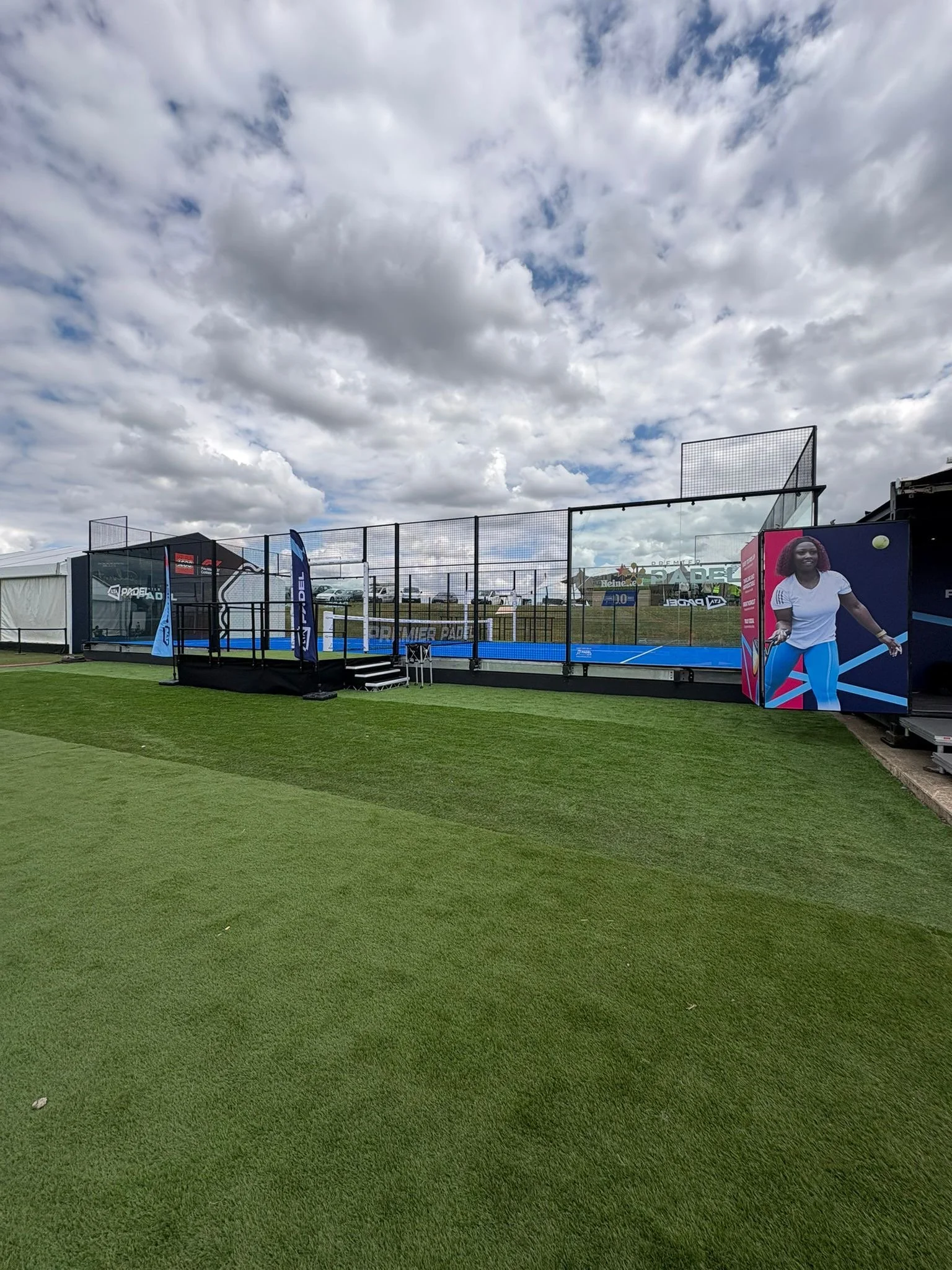 An outdoor Padel court with a blue surface, surrounded by black fencing, under a cloudy sky inside Silverstone Circuit. There are colourful banners.