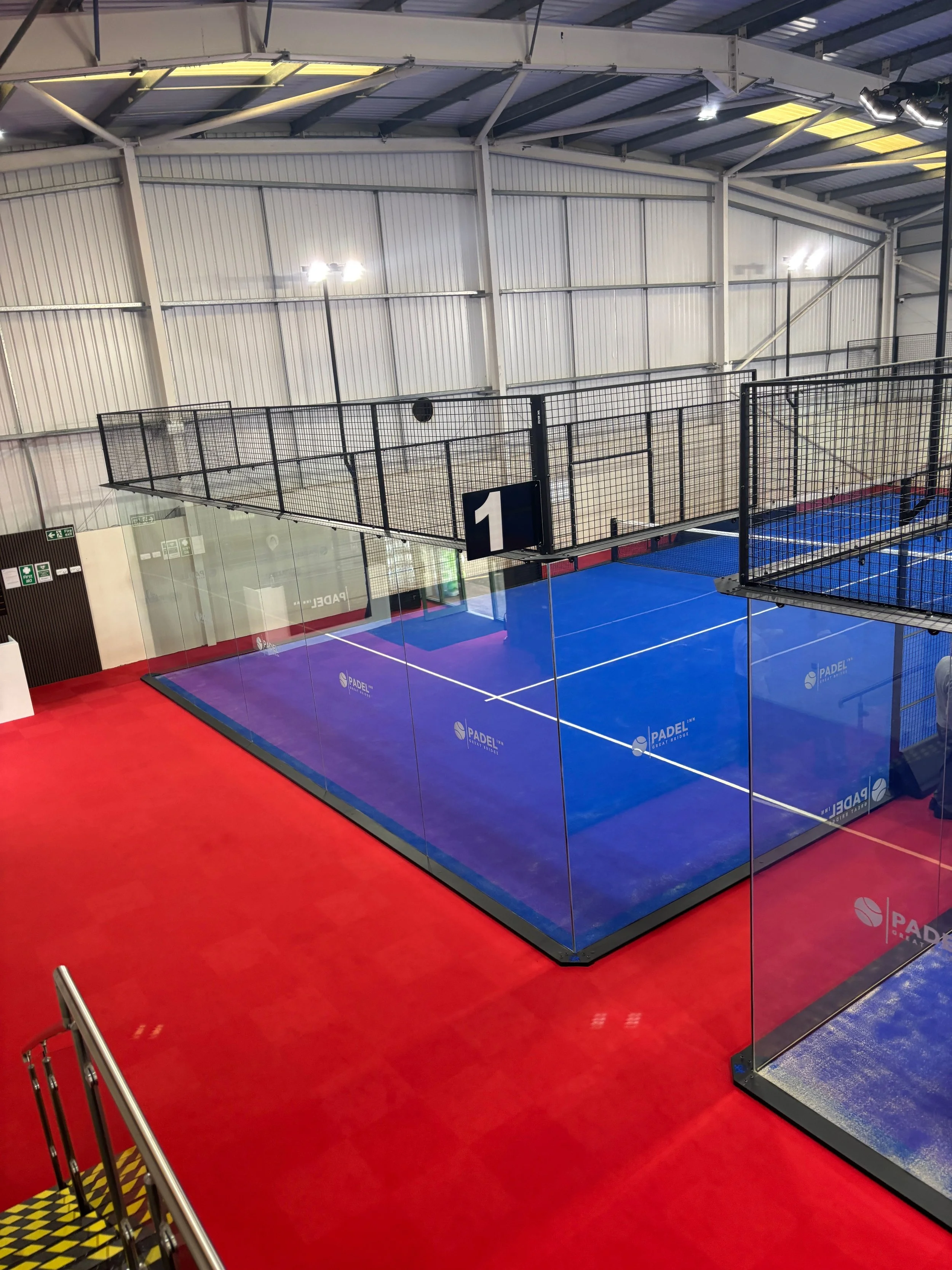 Indoor padel tennis court with blue playing surface surrounded by glass walls and black fencing UKs Best Padel