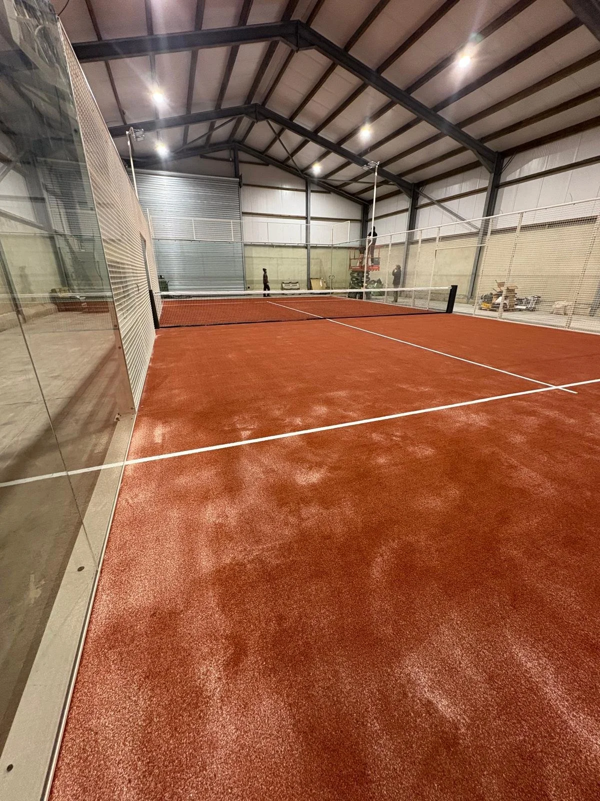 Indoor Padel court with red surface, surrounded by glass walls and metal structure, with people practicing in the background.