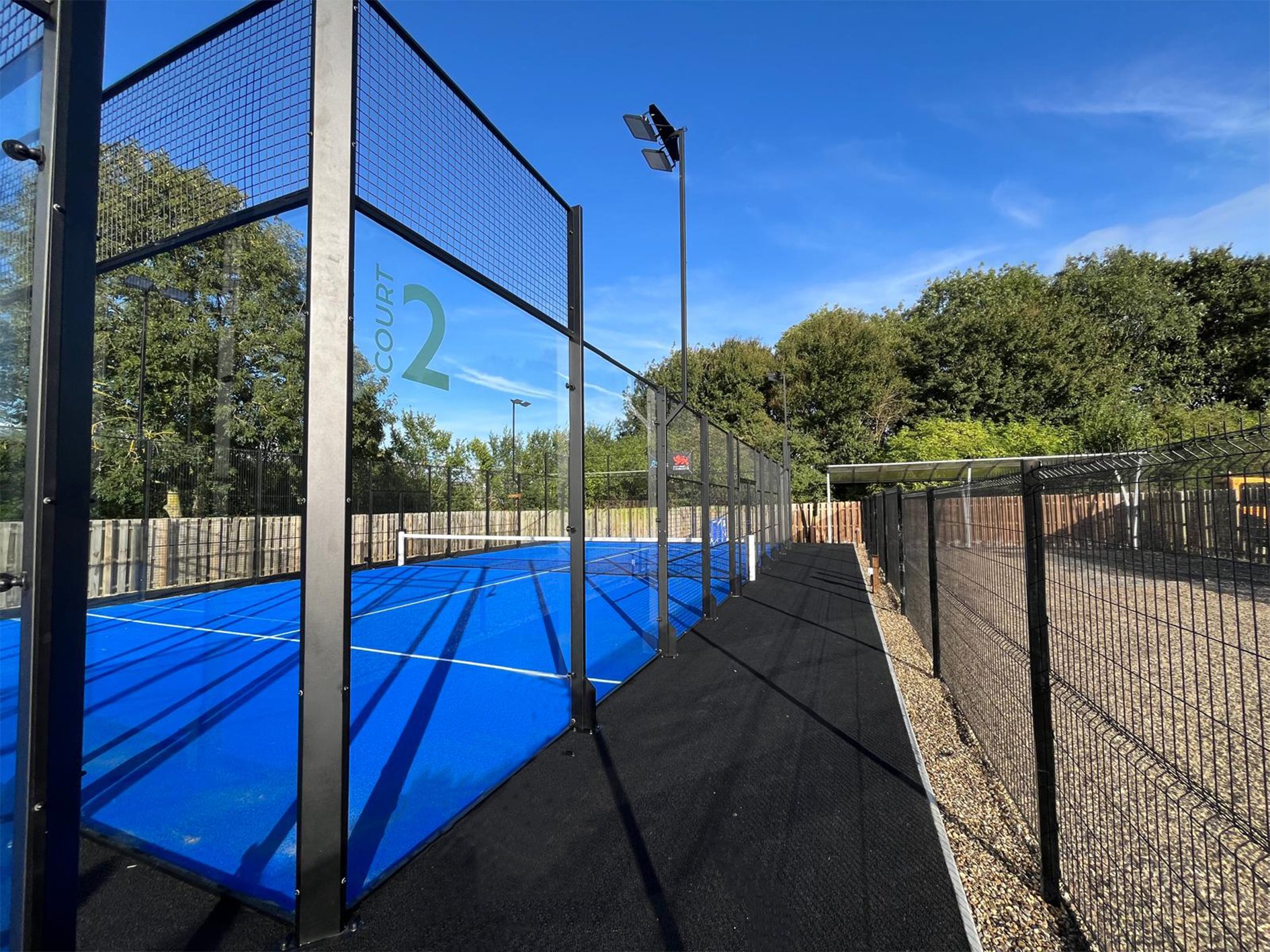 Padel court with blue surface, surrounded by fencing, under a clear blue sky with trees in the background and a sign that reads 'COURT 2'. University of Cambridge