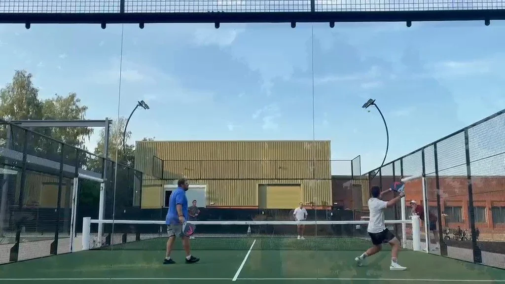 People playing padel tennis on an outdoor court with glass walls and a yellow building in the background at JCB golf and country club