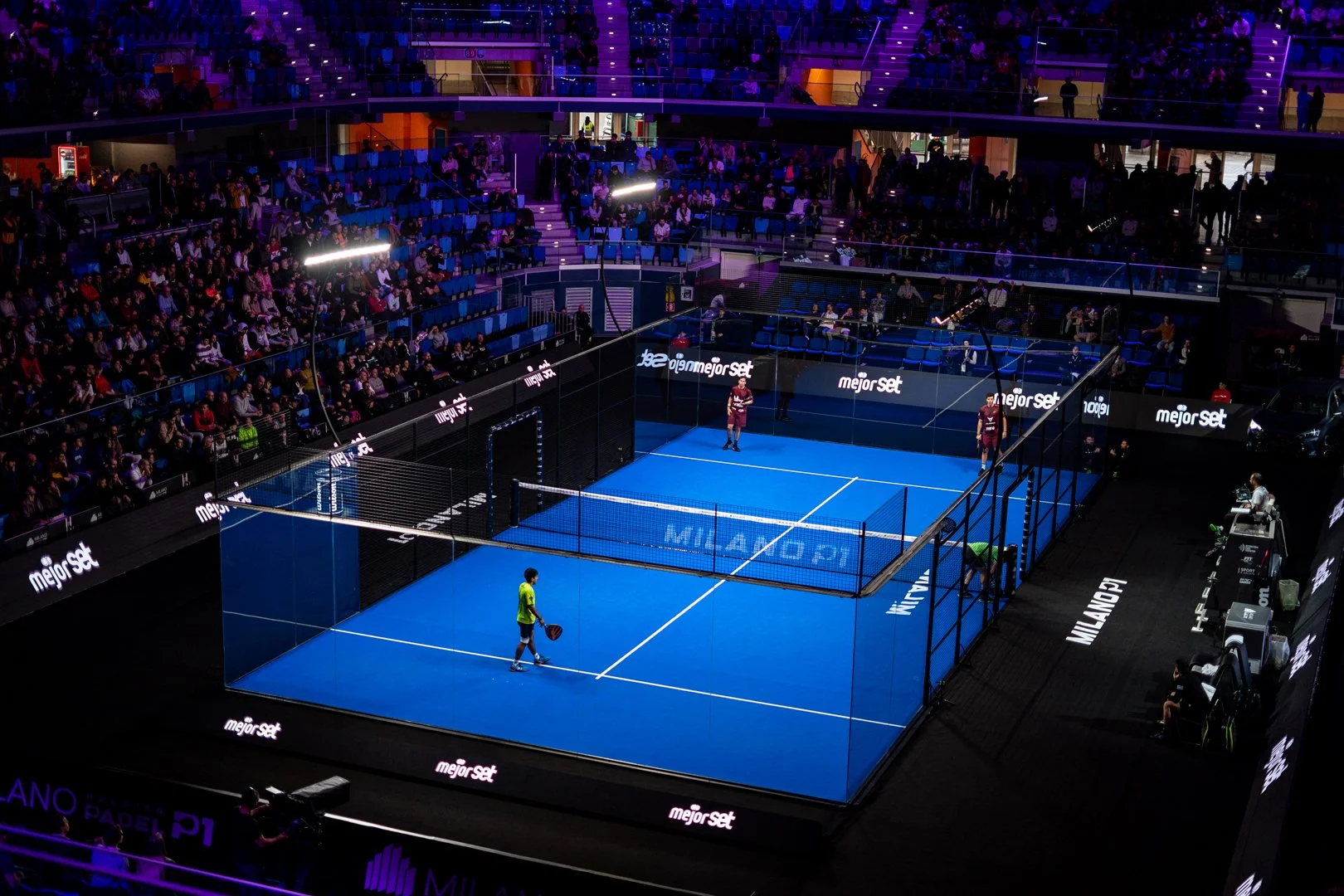 Indoor padel court with players and audience, lit by purple and white lighting, surrounded by advertising banners in a tournament setting.