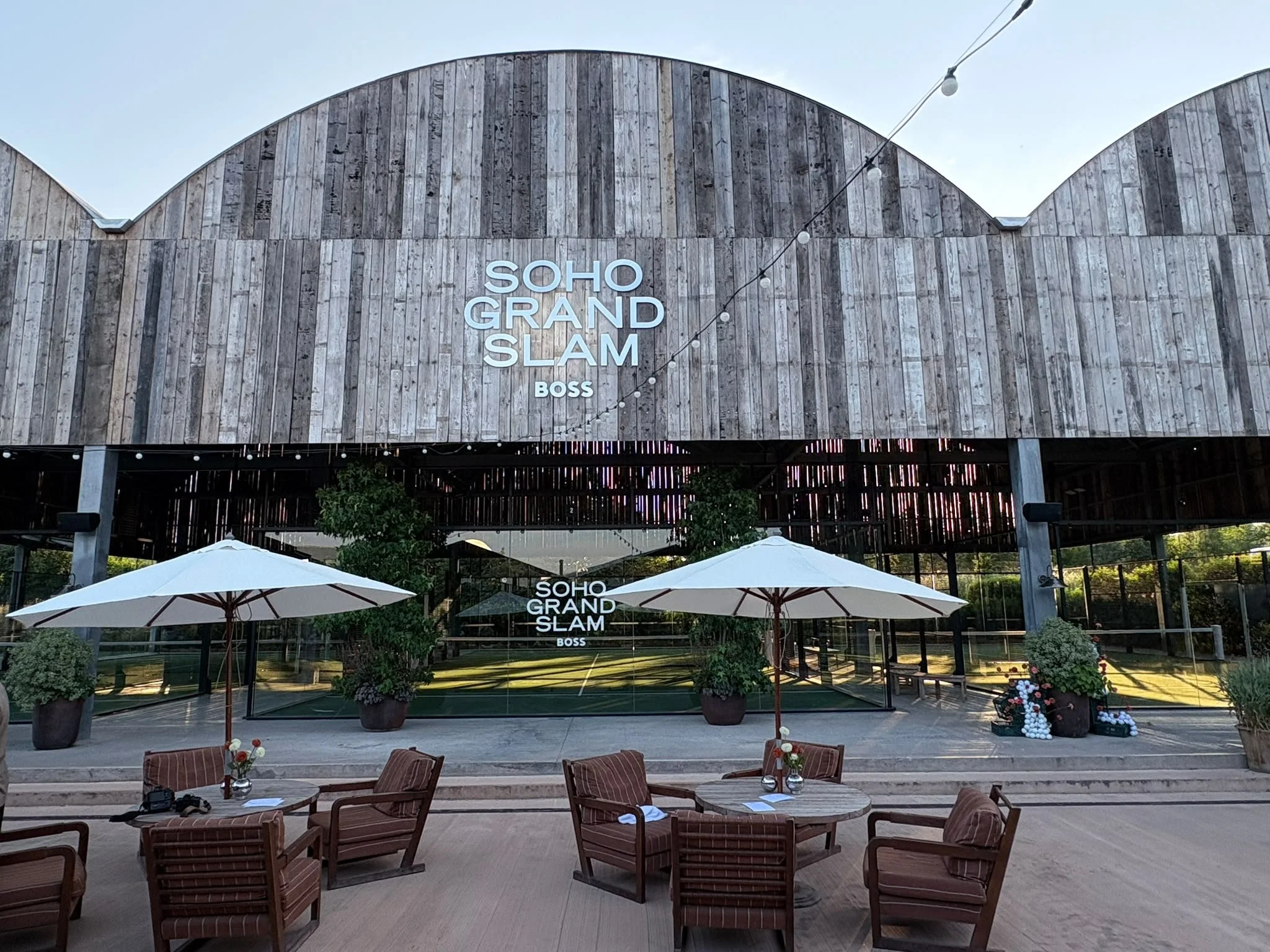 Outdoor venue with wooden Canopy labeled 'SOHO GRAND SLAM" with Padel Courts inside potted plants, umbrellas, and seating area.