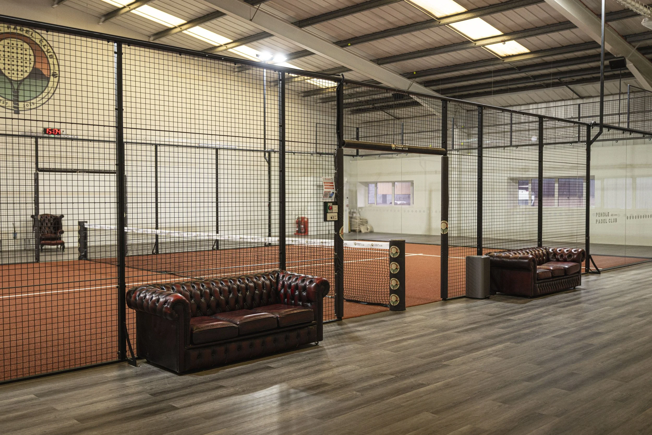 Indoor Terracotta padel court surrounded by black metal fencing, with leather couches and wood flooring.
