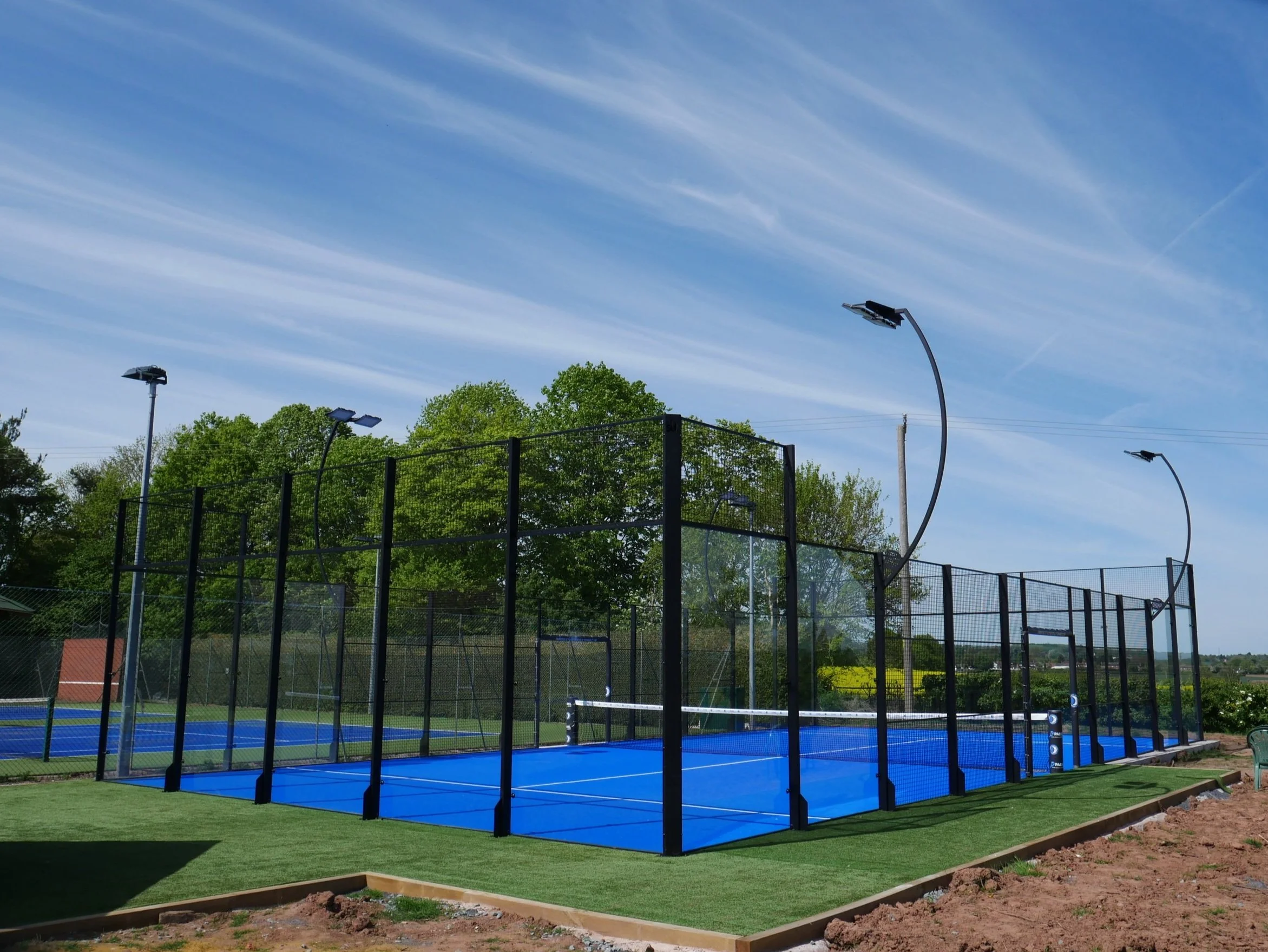 An outdoor pickleball court with blue surface, black fencing, and tall curved lights under a partly cloudy sky, surrounded by green trees.