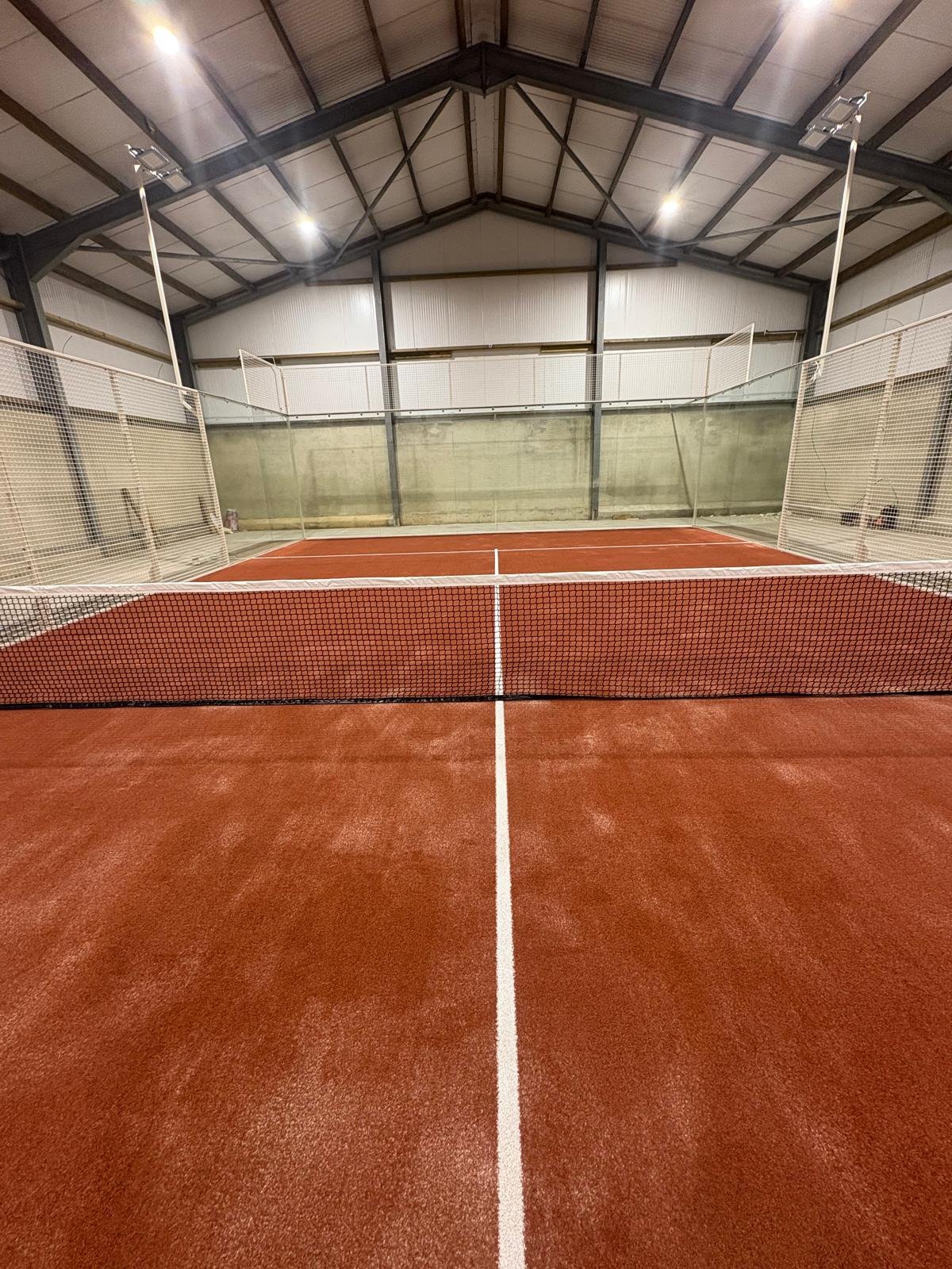 Indoor Padel court with red surface, white lines, net in the foreground, surrounded by high white walls, with a high ceiling and bright overhead lighting.